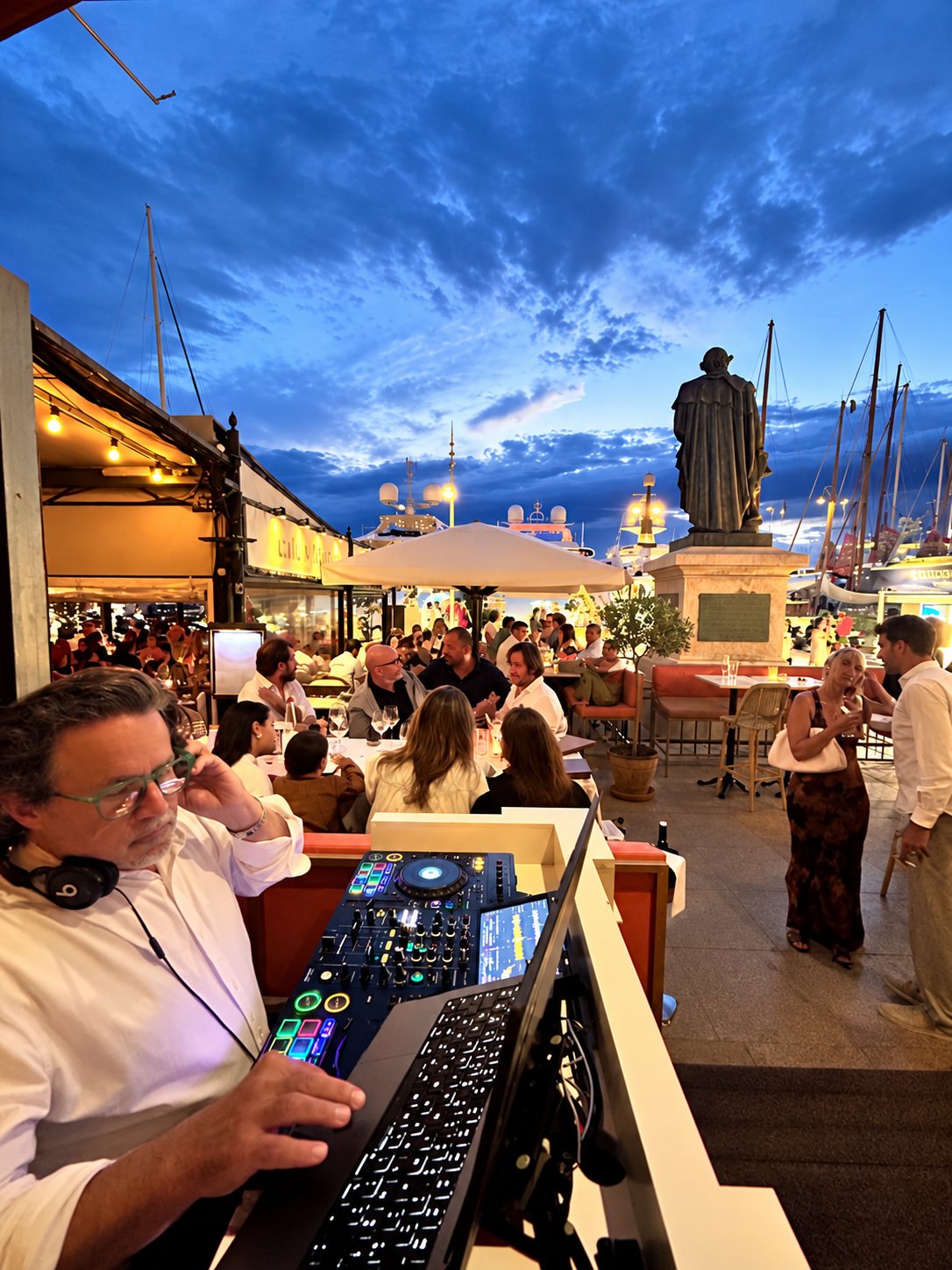 DJ aux platines sur une terrasse de restaurant face au port, au crépuscule.