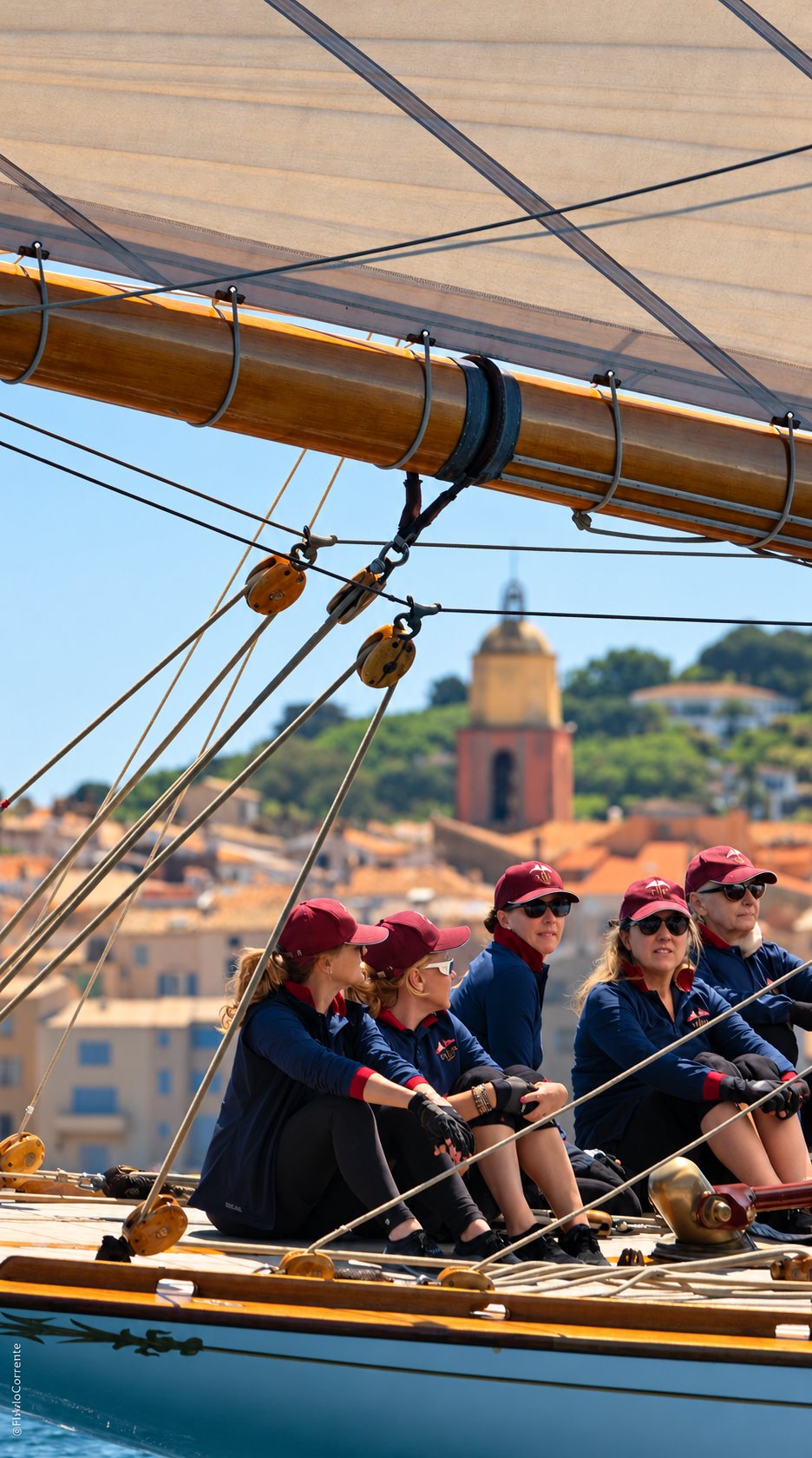 Cinq équipières en polos bleu marine et casquettes bordeaux assises sur le pont d’un voilier classique, devant un village côtier.