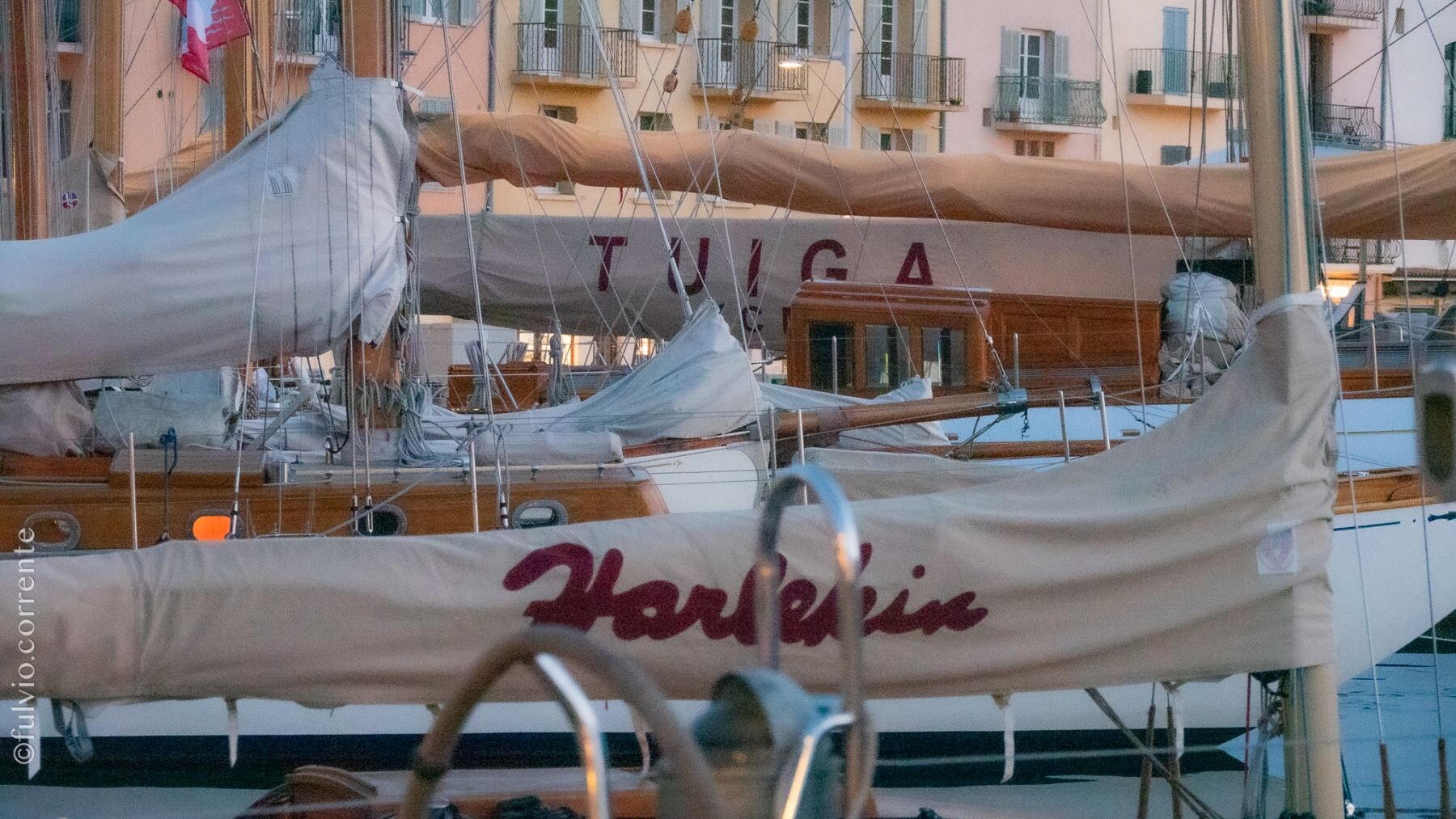 Voiliers au port, noms peints sur les housses de voile, façades colorées derrière.