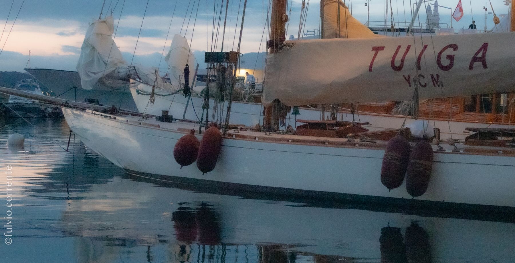 Coque blanche d’un voilier le long du quai, pare-battage rouges et reflets.