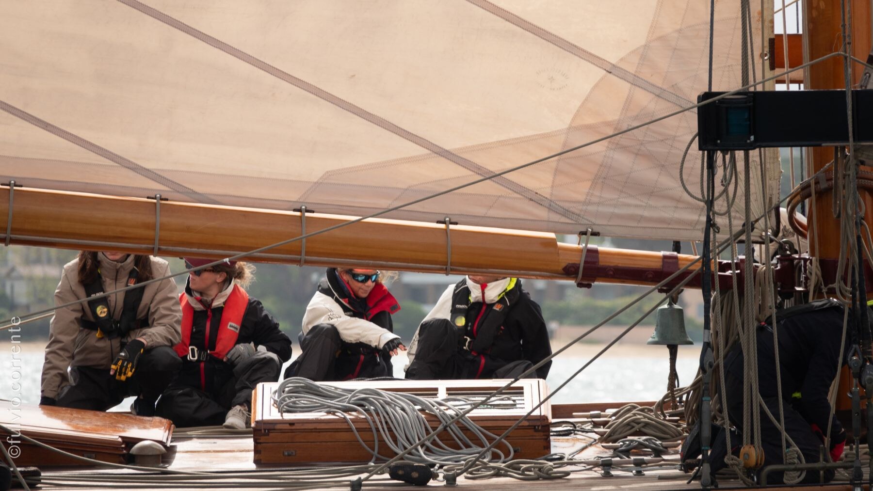 Équipiers en gilets assis sur le pont, sous la grande voile d’un voilier.