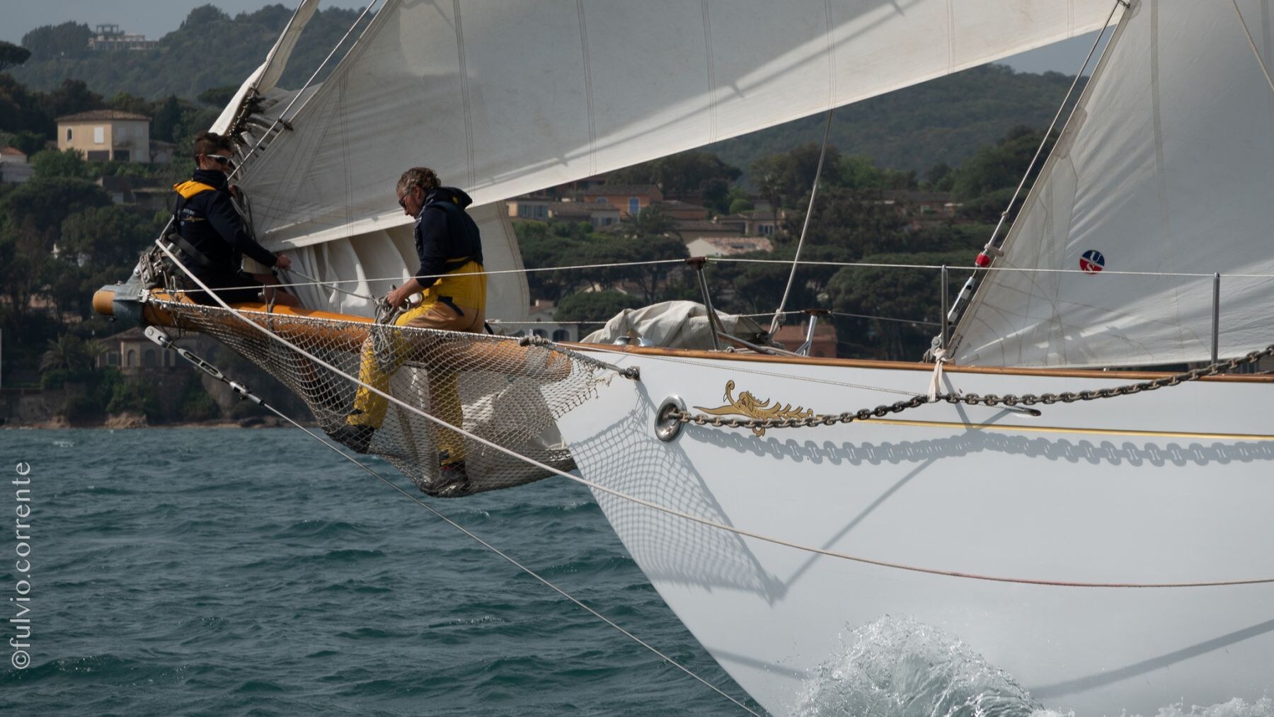 Deux marins sur le bout-dehors d’un grand voilier blanc, côte boisée derrière.