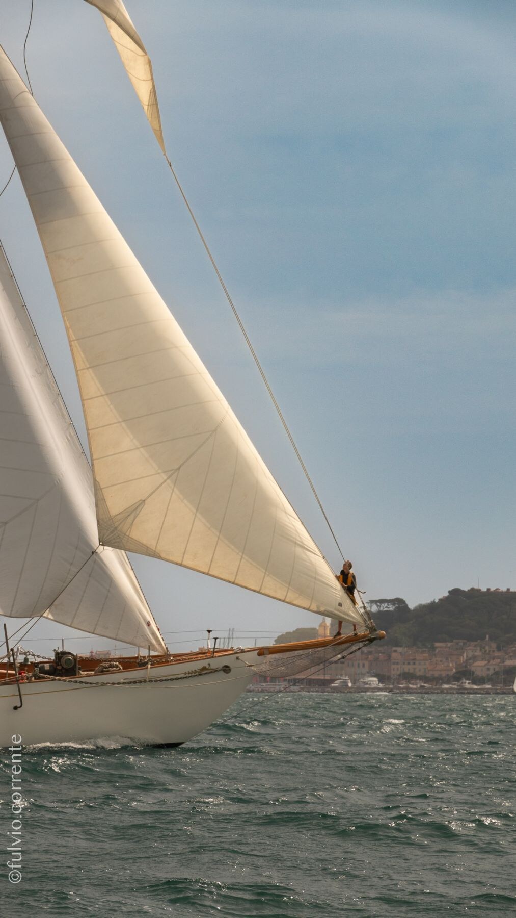 Voilier classique à voiles beiges, étrave blanche, côte rocheuse.