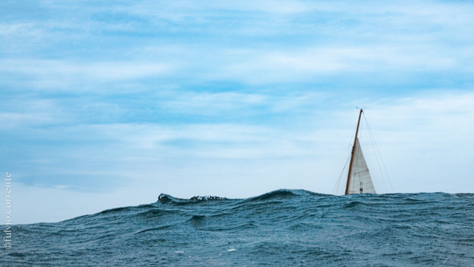 Petite voile blanche au loin derrière une vague, ciel bleu.