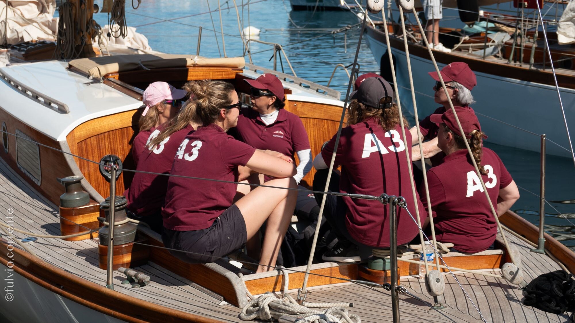 Équipières en polos bordeaux numérotés, assises sur le pont d’un voilier classique au port.