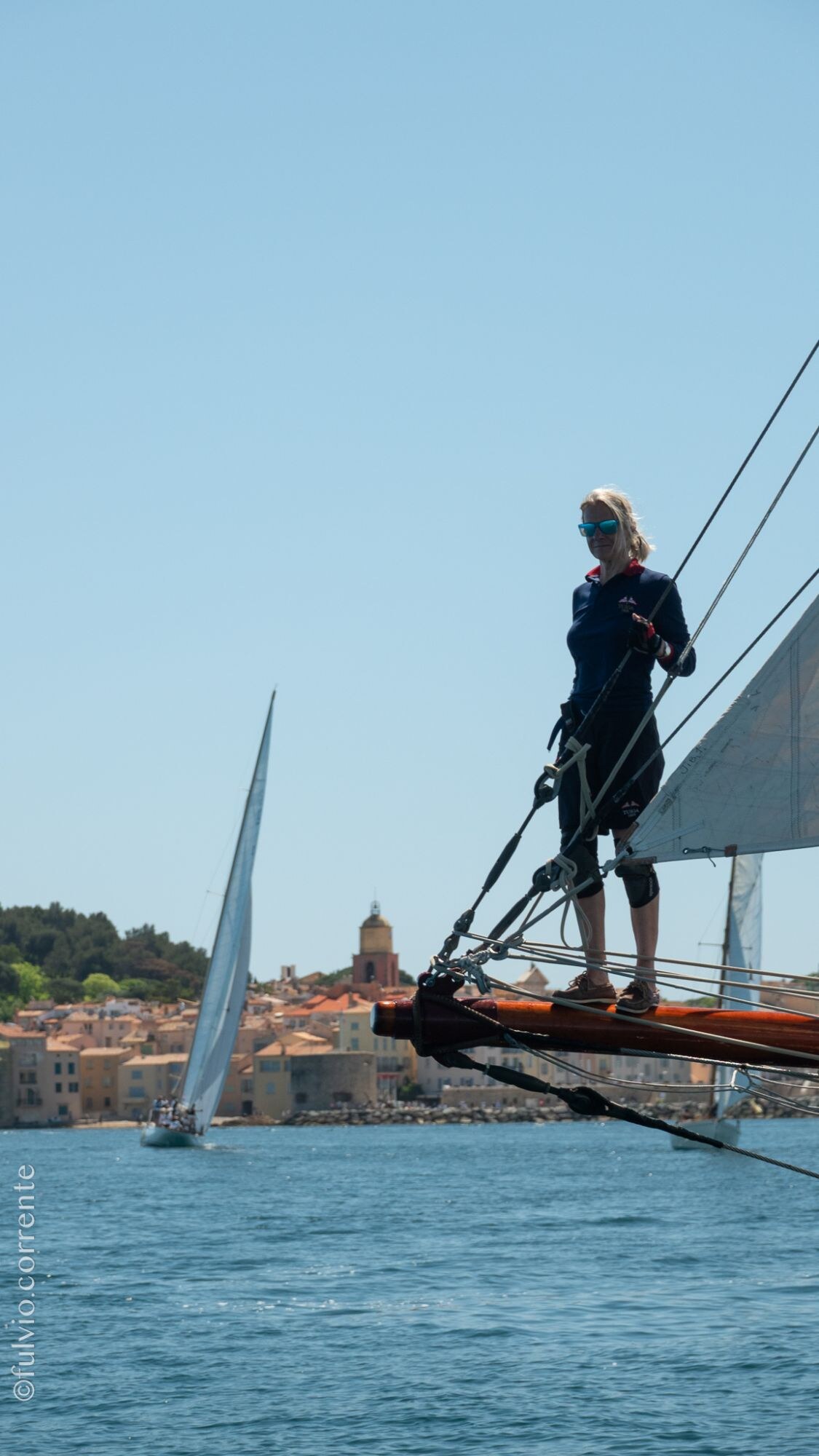 Équipière debout sur le bout-dehors d’un voilier, village de Saint-Tropez au loin.