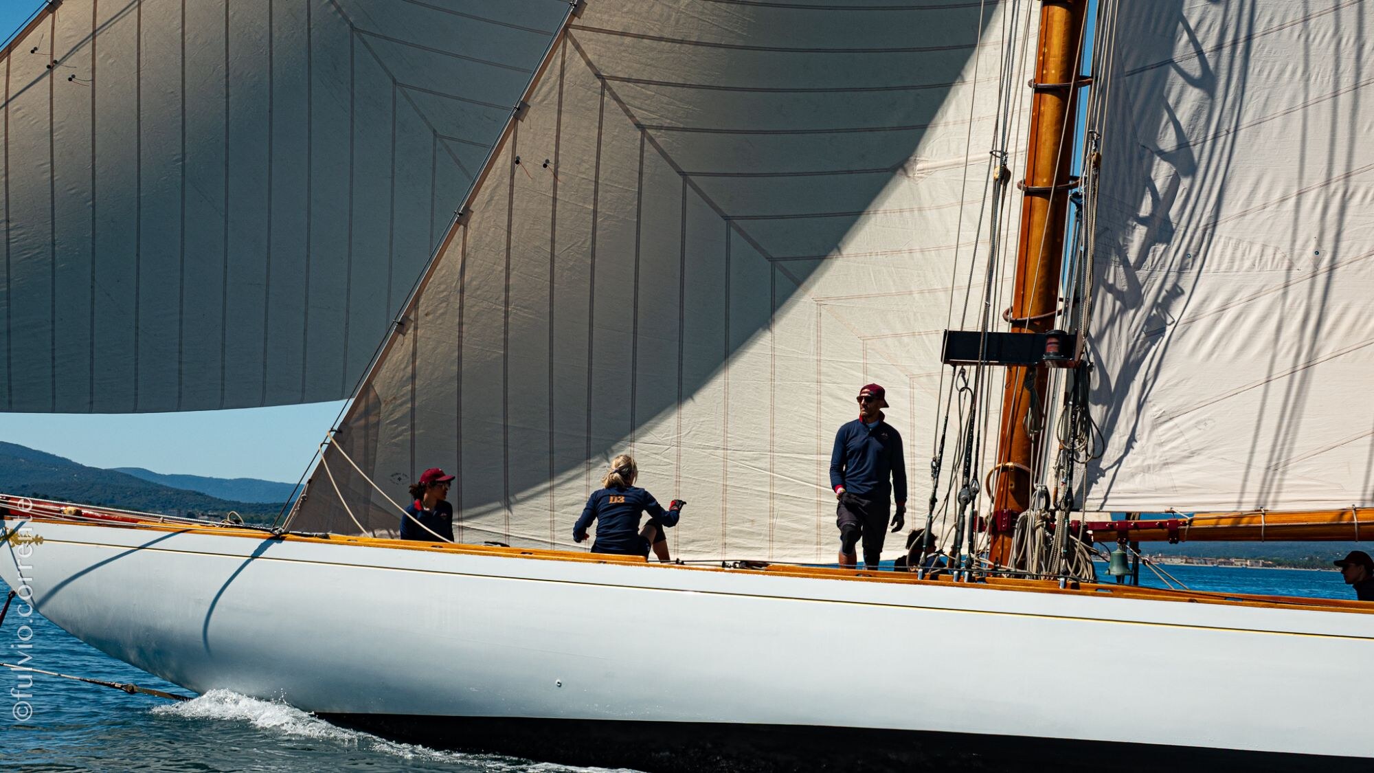 Équipiers sur le pont d’un voilier blanc sous ses grandes voiles, vus de l’avant.