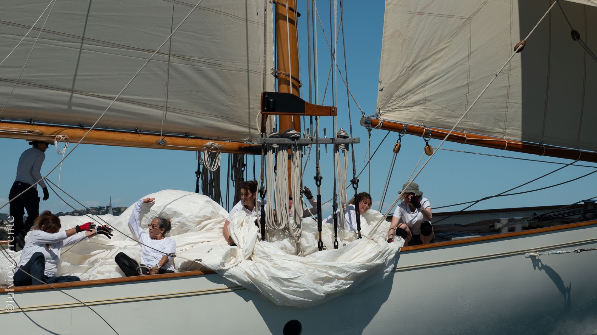 Équipières en blanc s’affairant dans la voile affalée sur le pont d’un voilier.