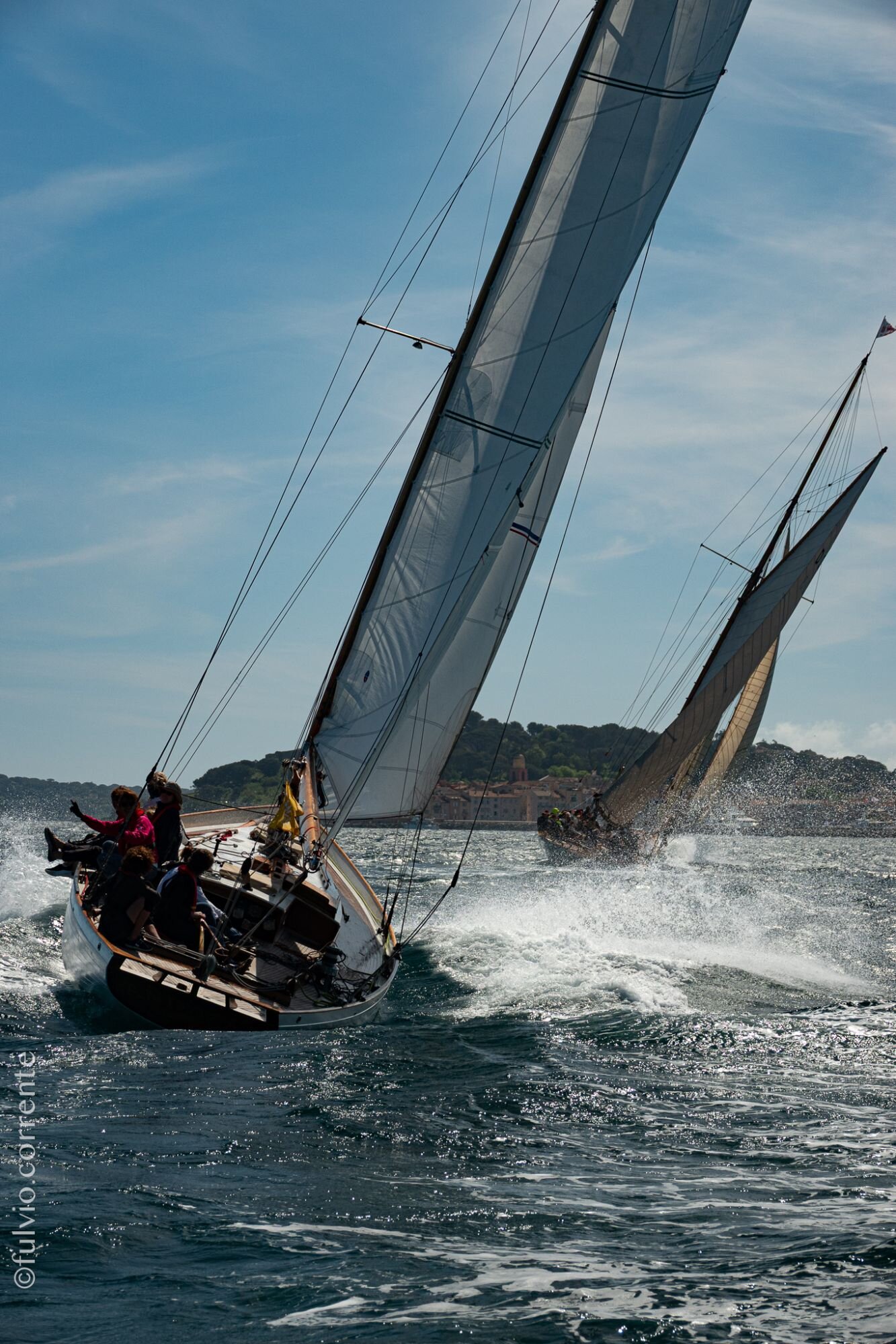 Voilier classique gîté fendant la mer, équipage penché et embruns, côte au fond.