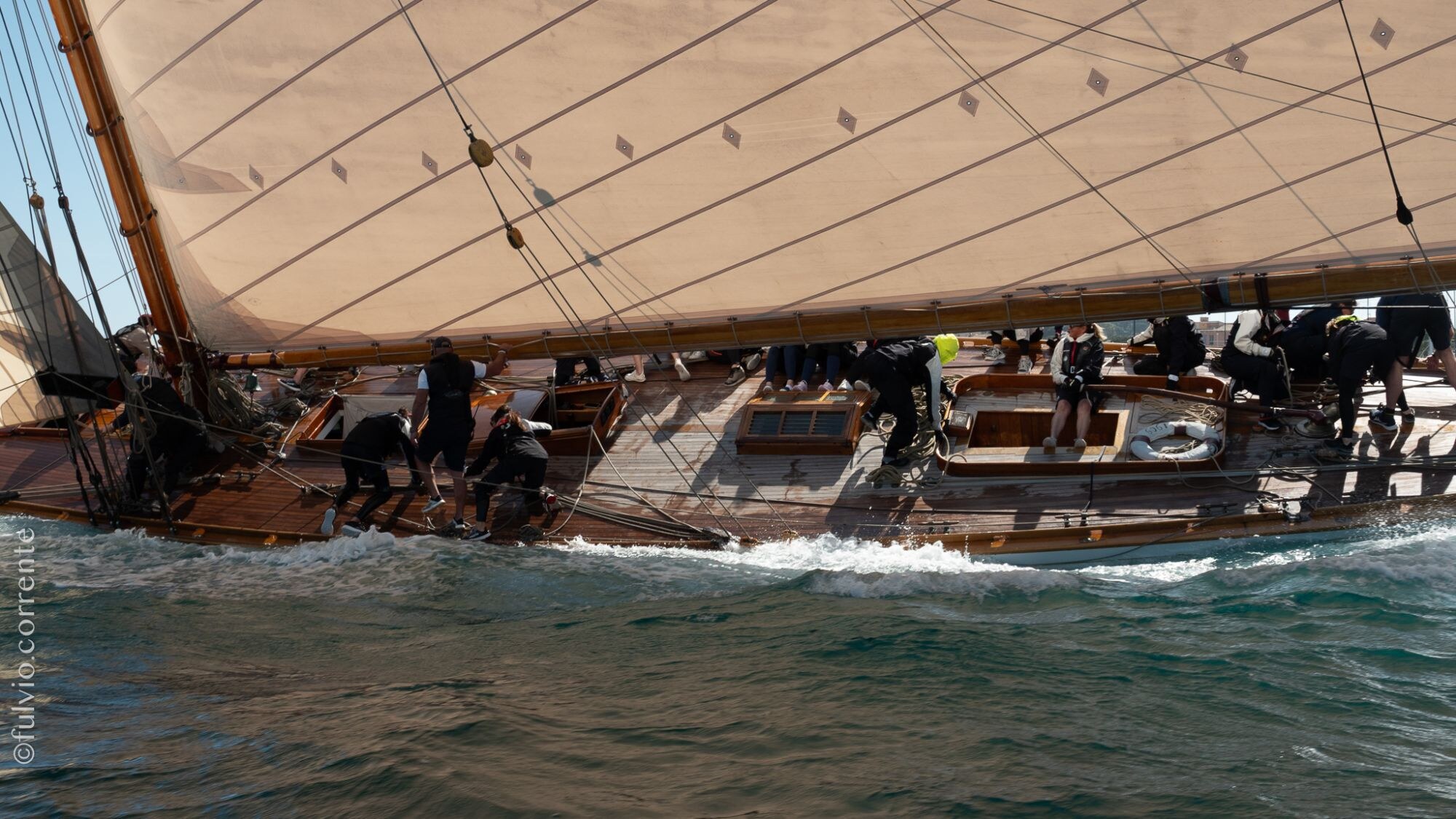 Équipage au travail sous la grande voile beige d’un voilier classique, vagues au premier plan.