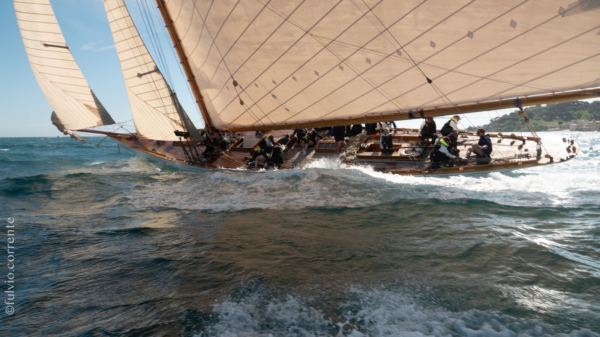 Voilier classique sous grande voile beige et deux focs, équipage sur le pont, gerbes d’écume.