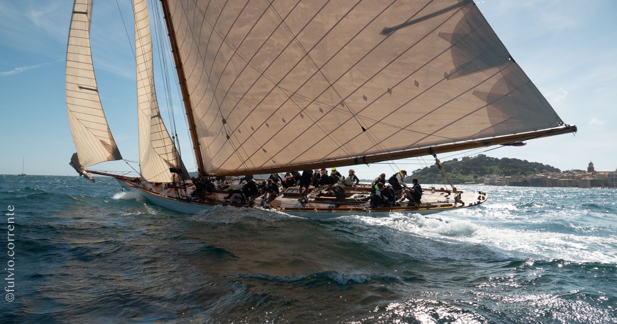 Voilier classique à grande voile brune sur une mer agitée, côte au fond.