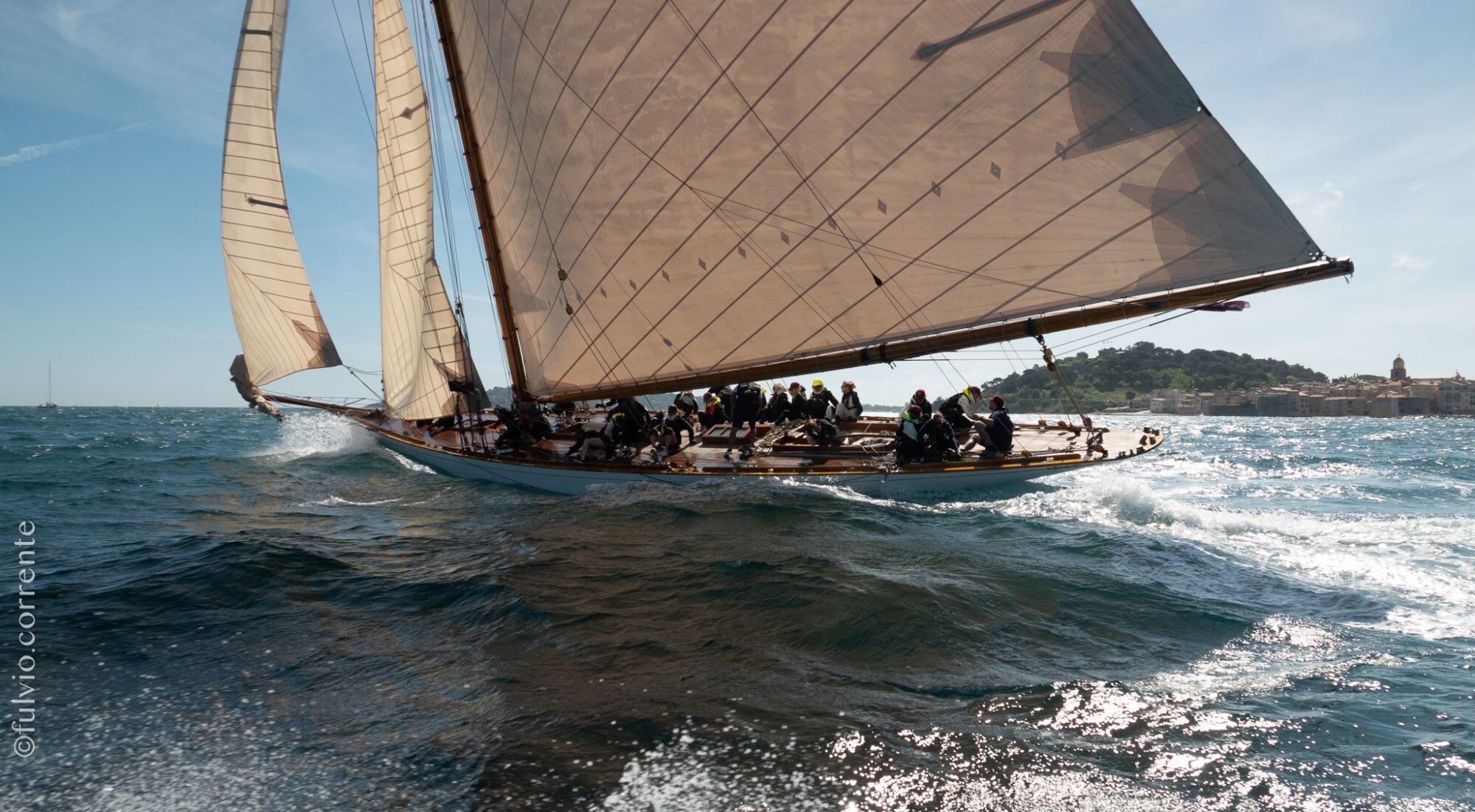 Voilier classique à grande voile brune, équipage assis au vent, vagues au premier plan.