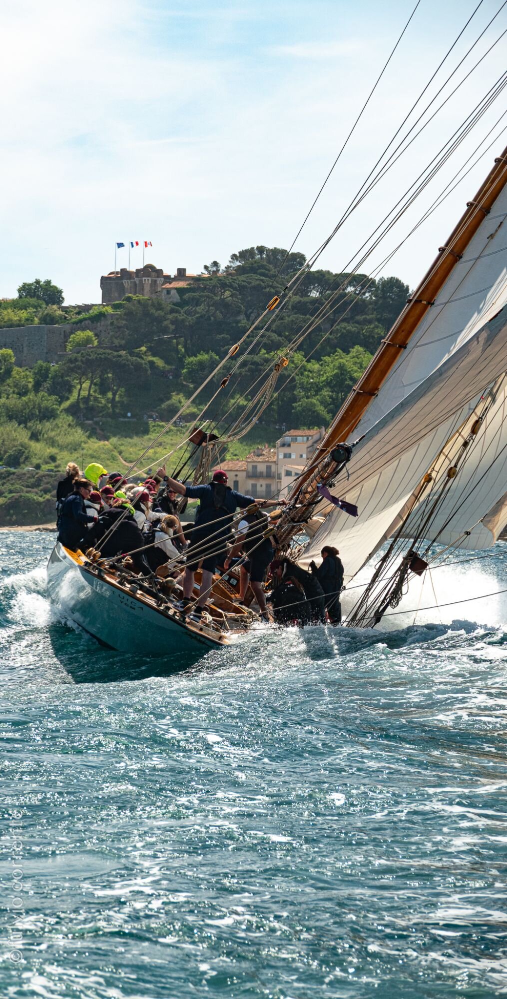 Voilier classique gîté devant une colline boisée et son fort, équipage au vent.