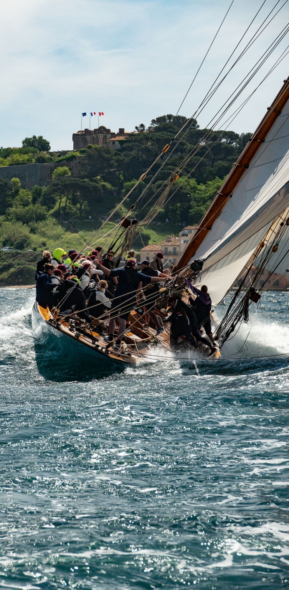 Voilier classique gîté devant la colline et son fort, équipage serré au vent.