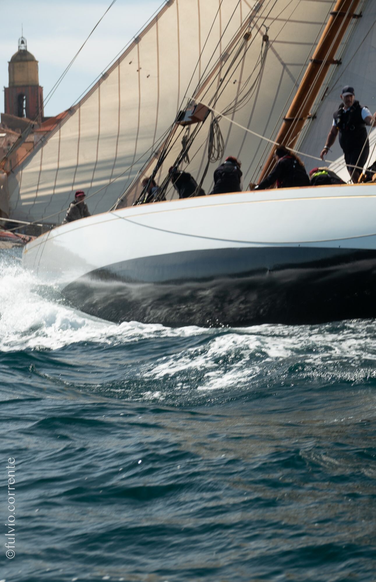 Coque blanche d’un voilier filant dans les vagues, clocher de Saint-Tropez à gauche.