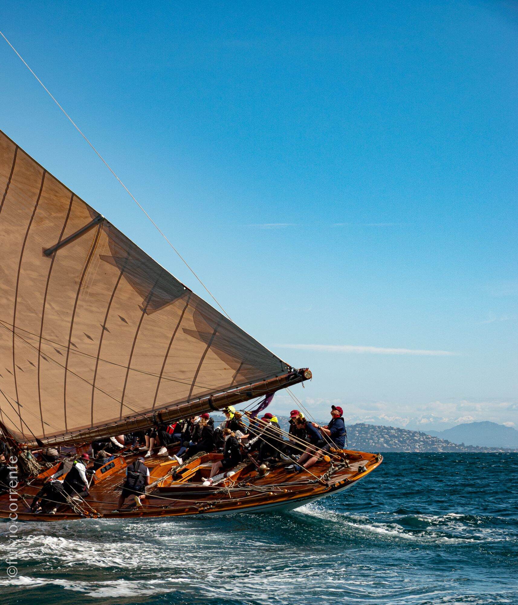 Étrave d’un voilier classique sous grande voile brune, côte lointaine et ciel bleu.