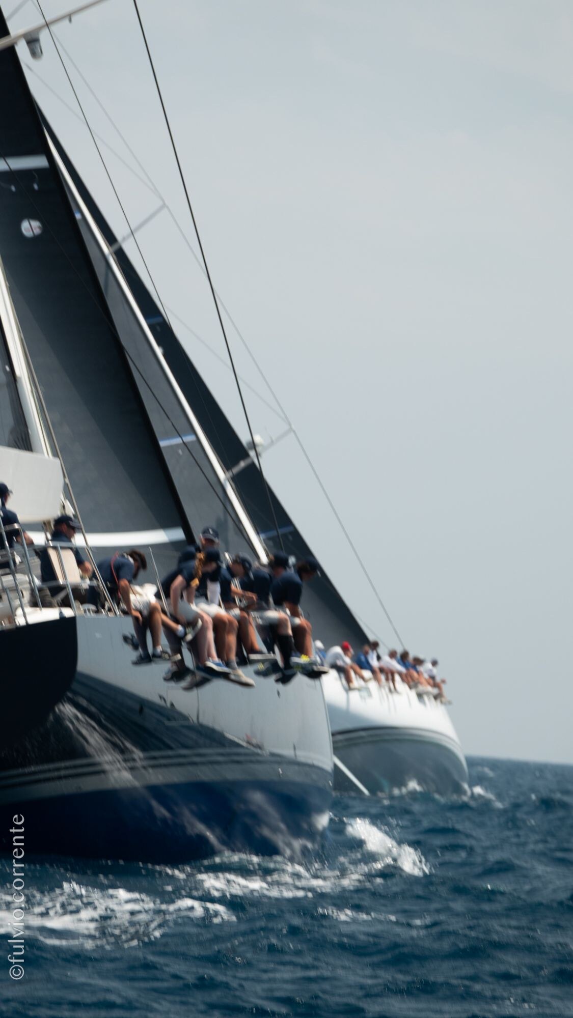 Équipage assis au rappel sur le bord d’un grand voilier de course à coque sombre.