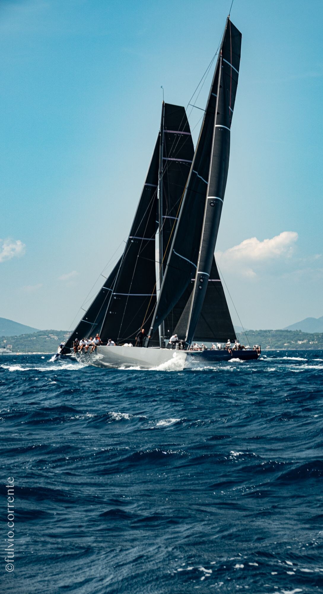 Voiliers de course aux voiles noires sous un ciel bleu, mer houleuse.