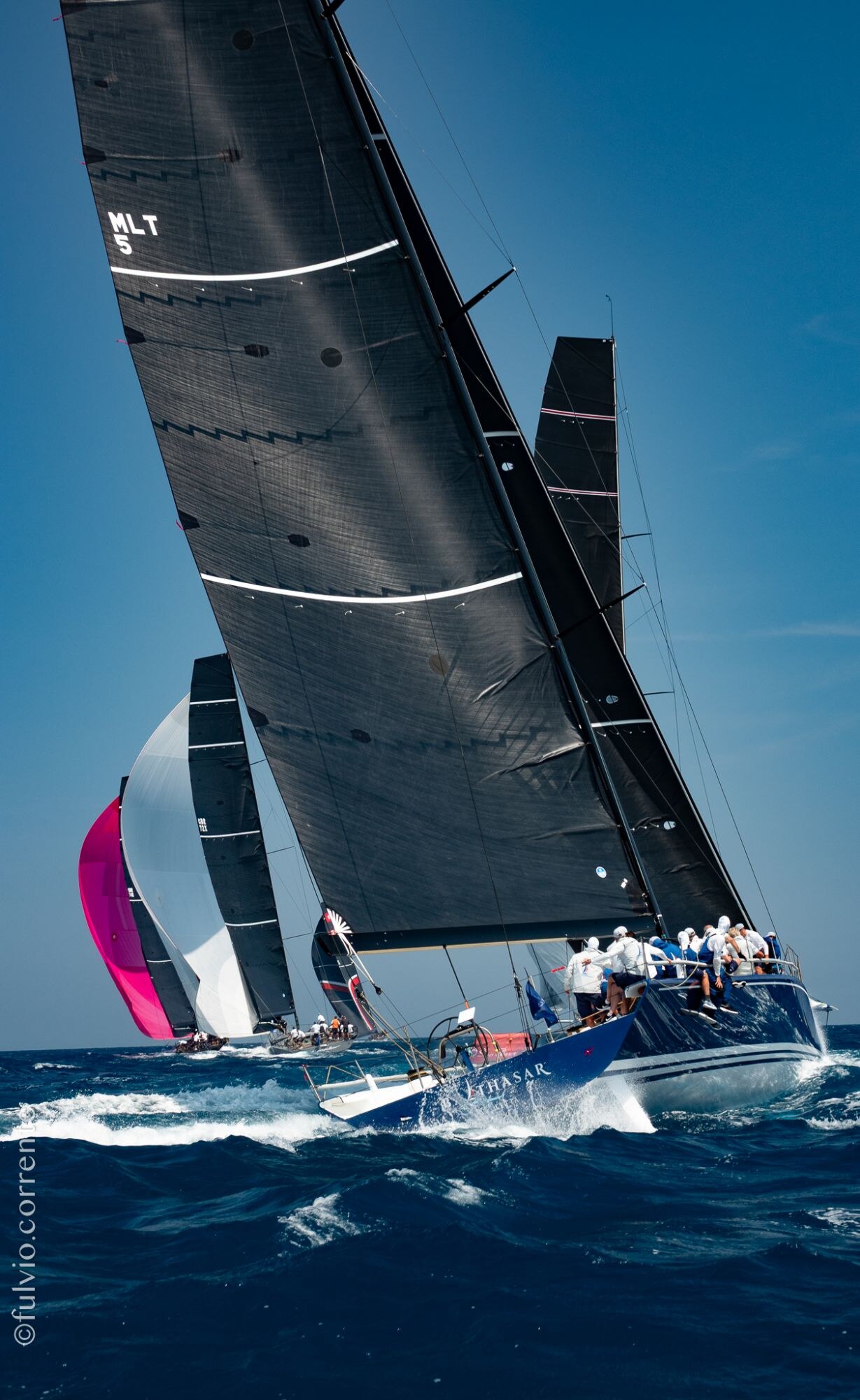 Grand voilier de course à grande voile noire gîté, spinnaker rose derrière.