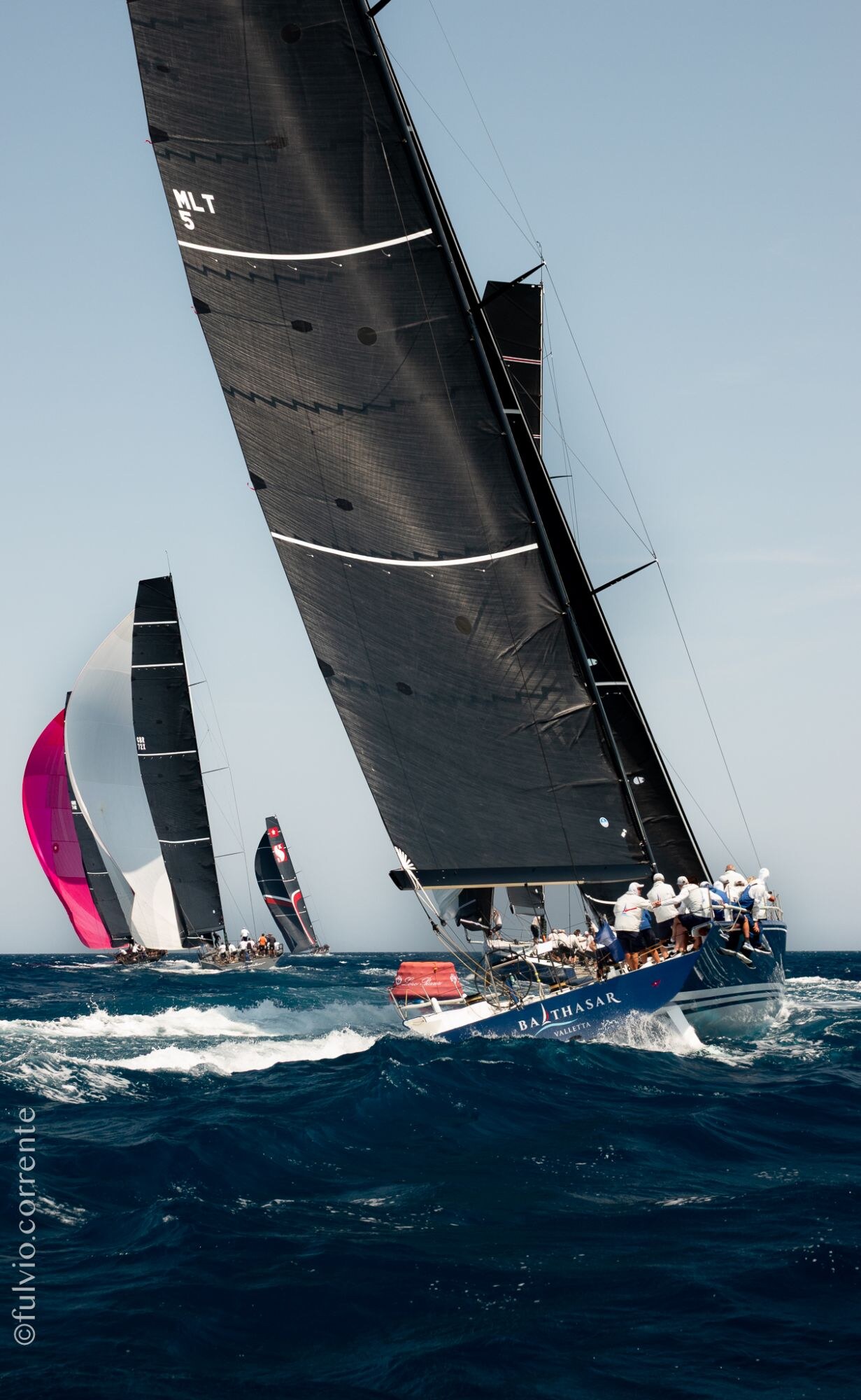 Voilier de course à voile noire gîté, spinnaker rose à l’arrière-plan.
