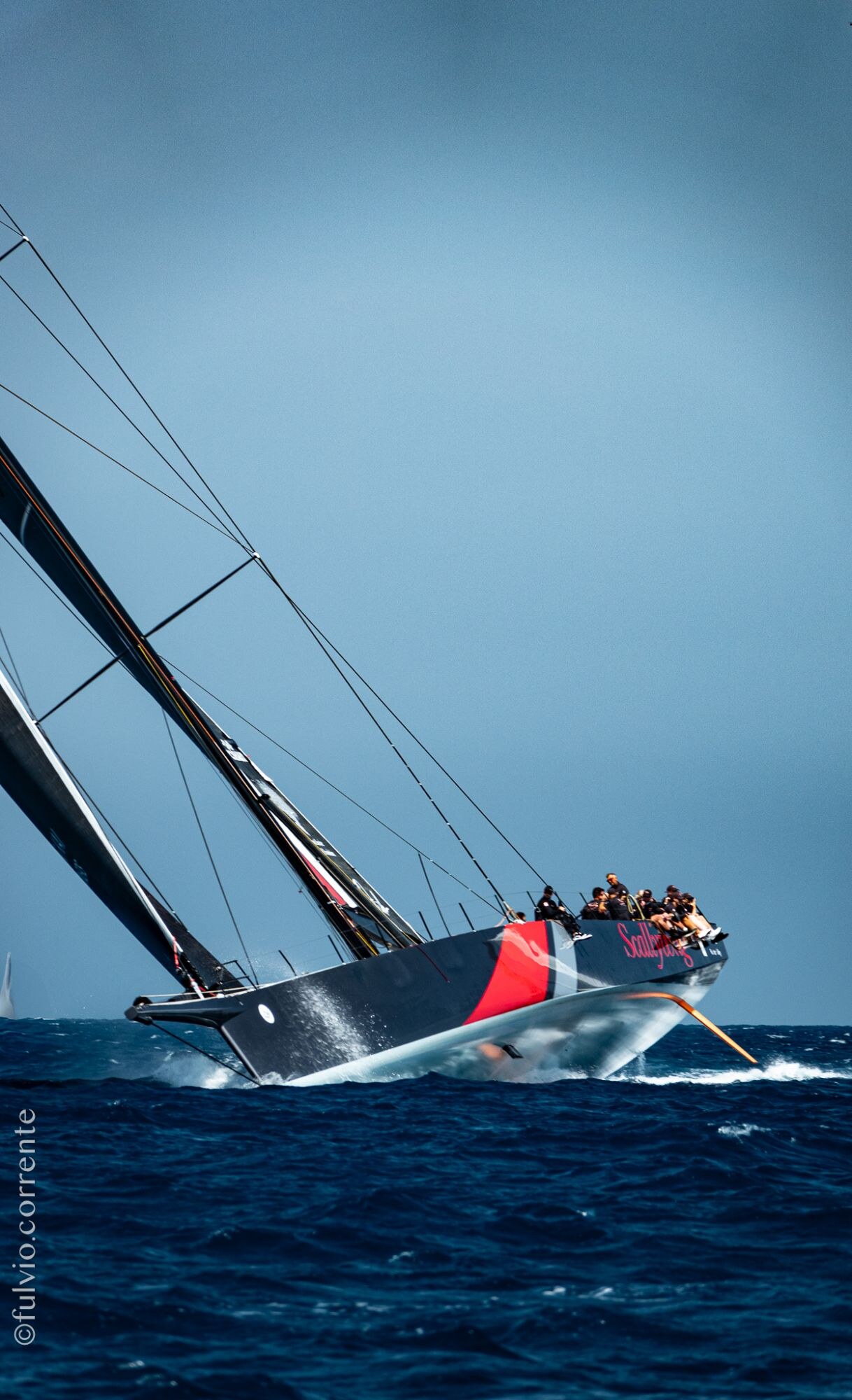 Voilier de course noir et rouge fortement gîté sur une mer bleu profond.