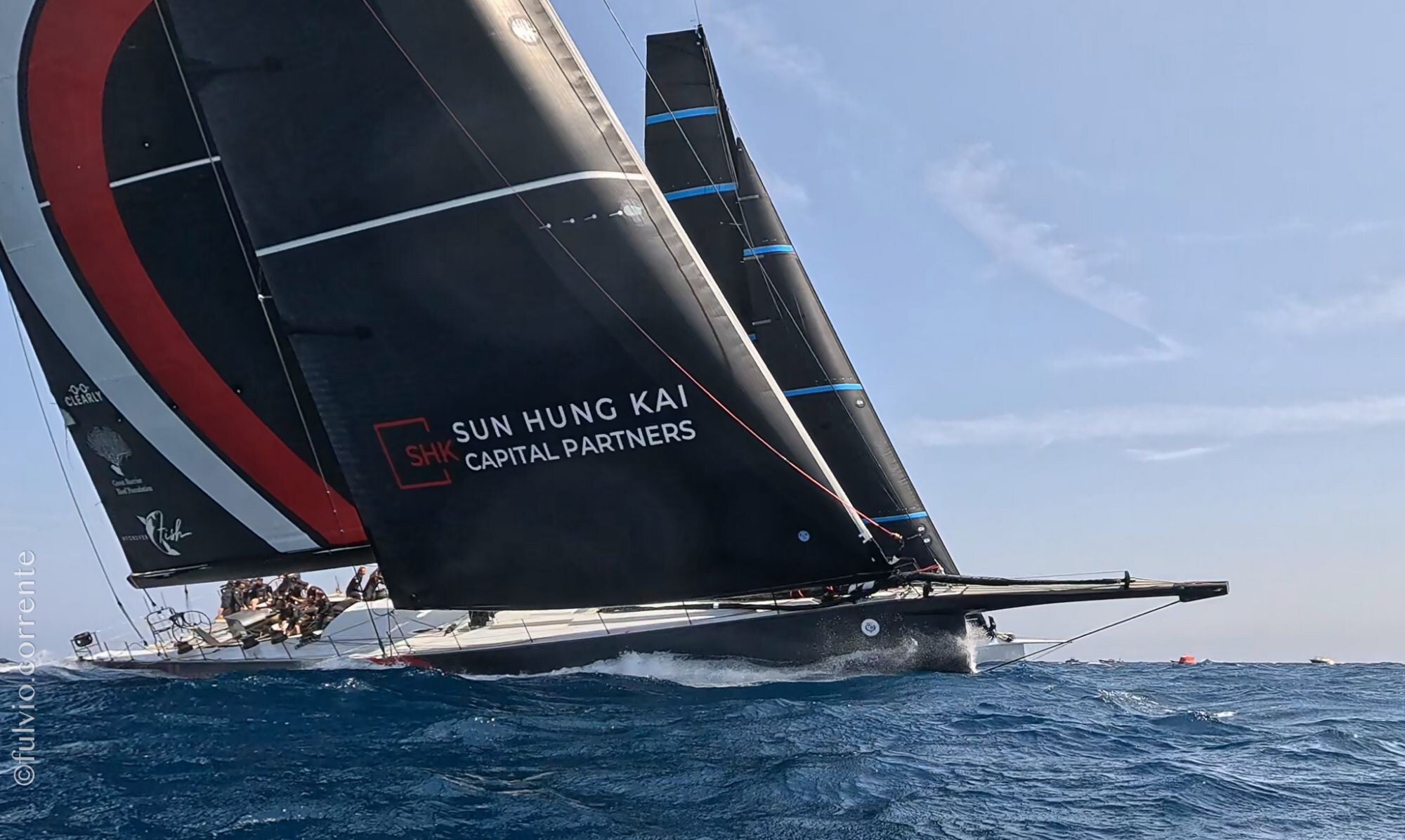 Grand voilier de course aux voiles noires et rouges portant un nom de sponsor.