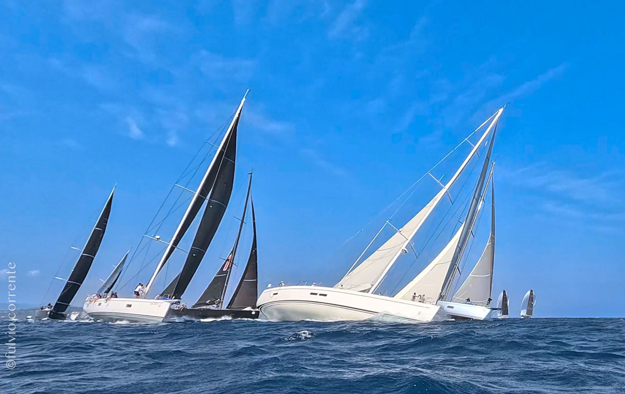 Voiliers de course aux voiles noires et blanches sur une mer bleue sous un ciel azur.
