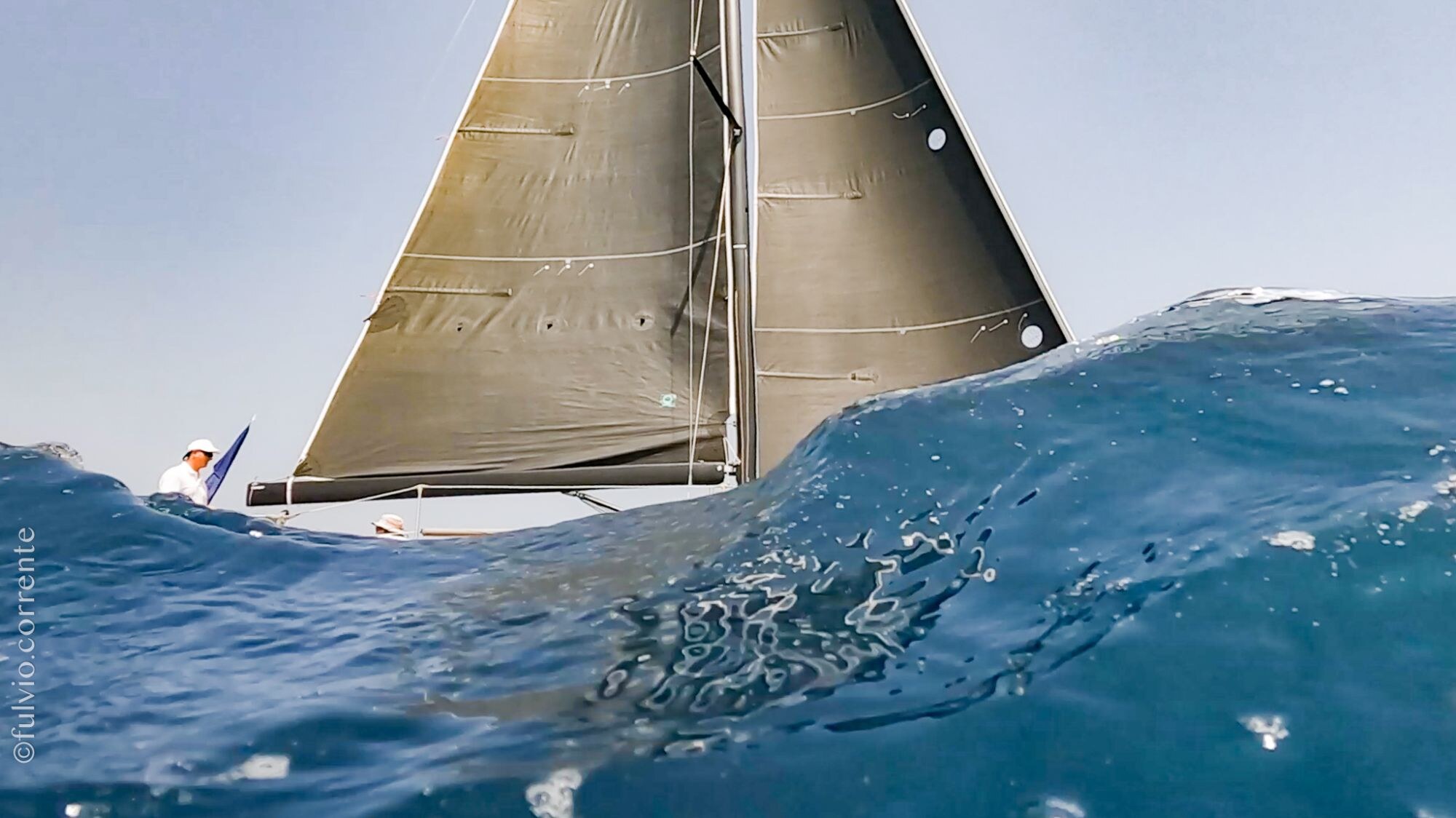 Vue basse d’un voilier à voiles ocre derrière une vague translucide.