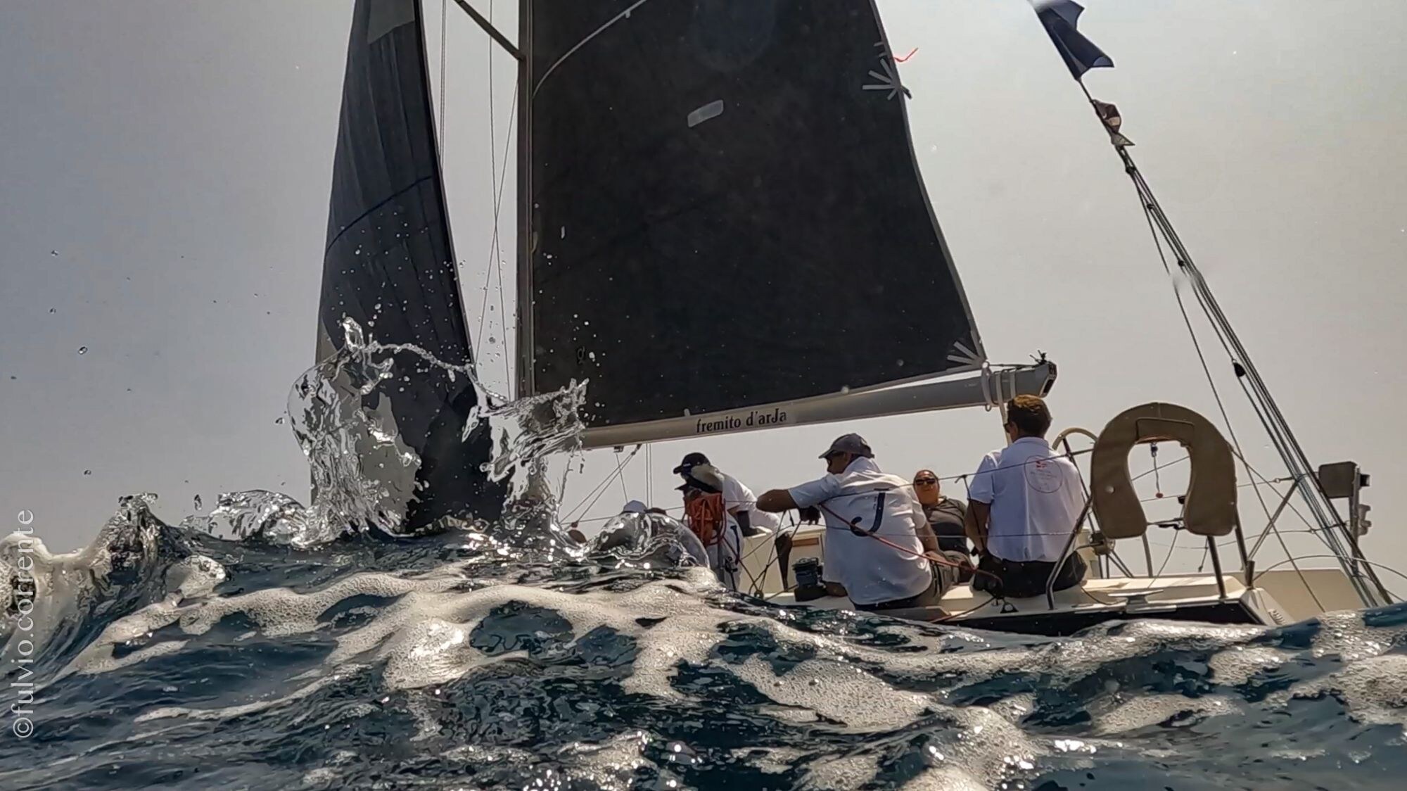 Équipage à l’arrière d’un voilier à voile noire, éclaboussures au premier plan.