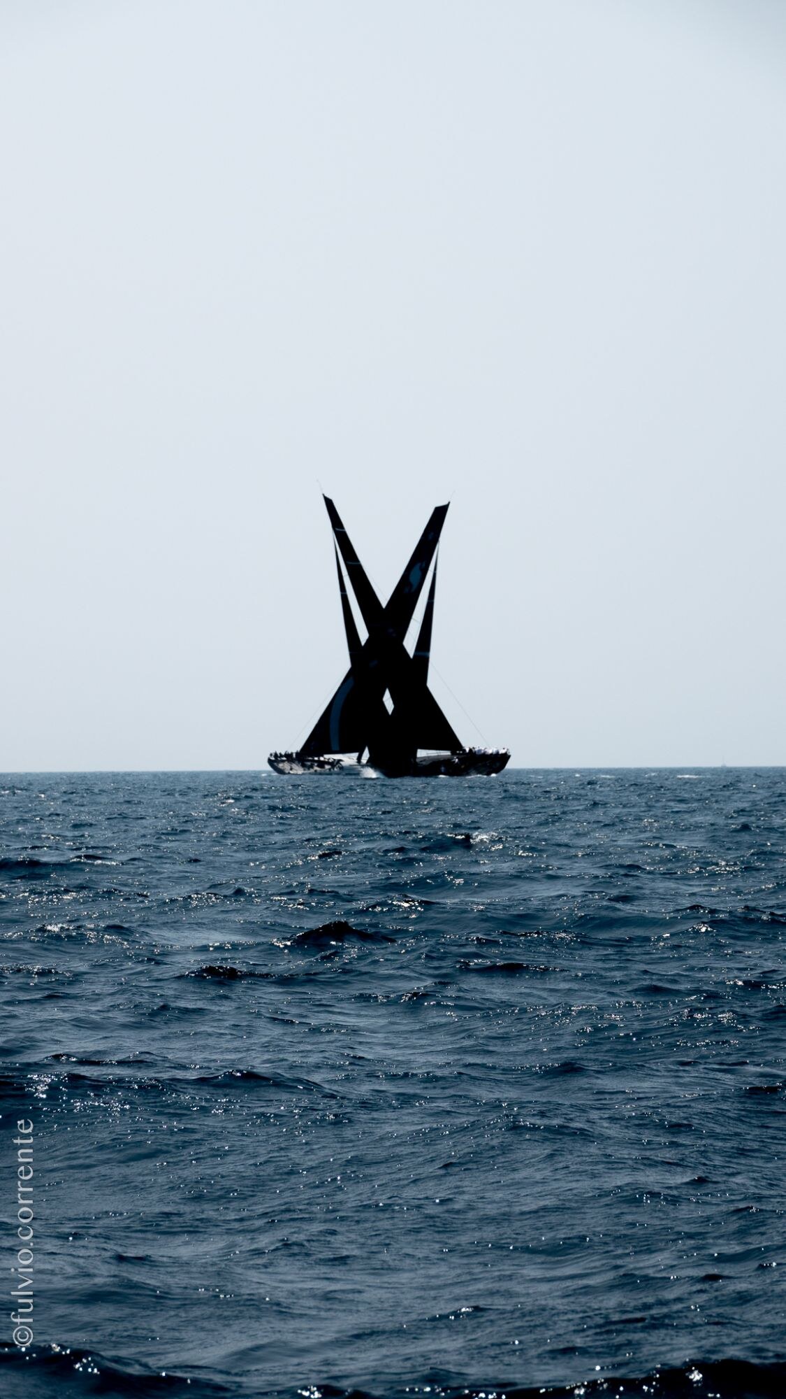 Silhouette sombre de voiliers de course à l’horizon sur une mer grise.