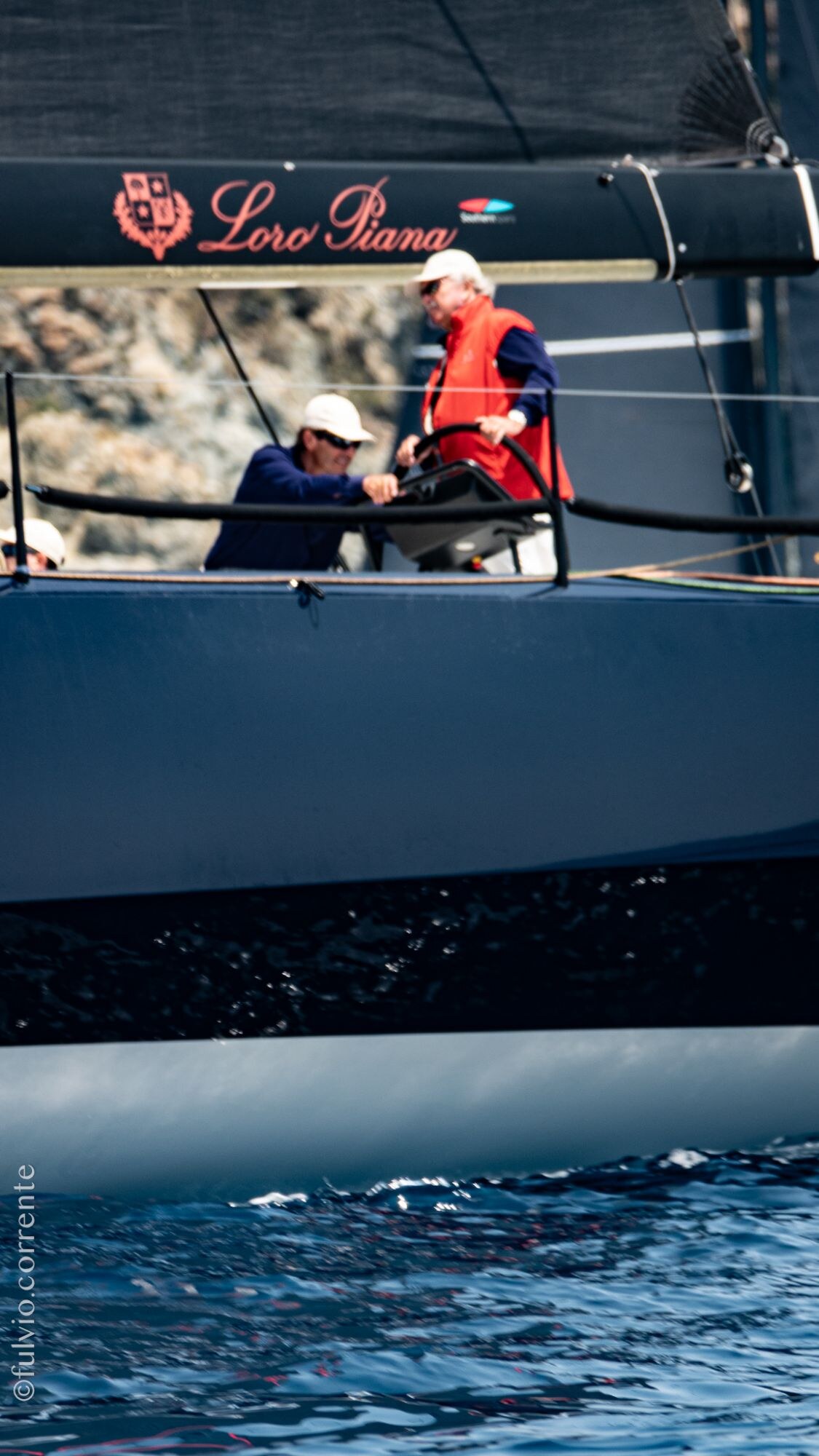 Équipiers en veste rouge sur le pont d’un voilier de course sombre, bandeau de marque en haut.
