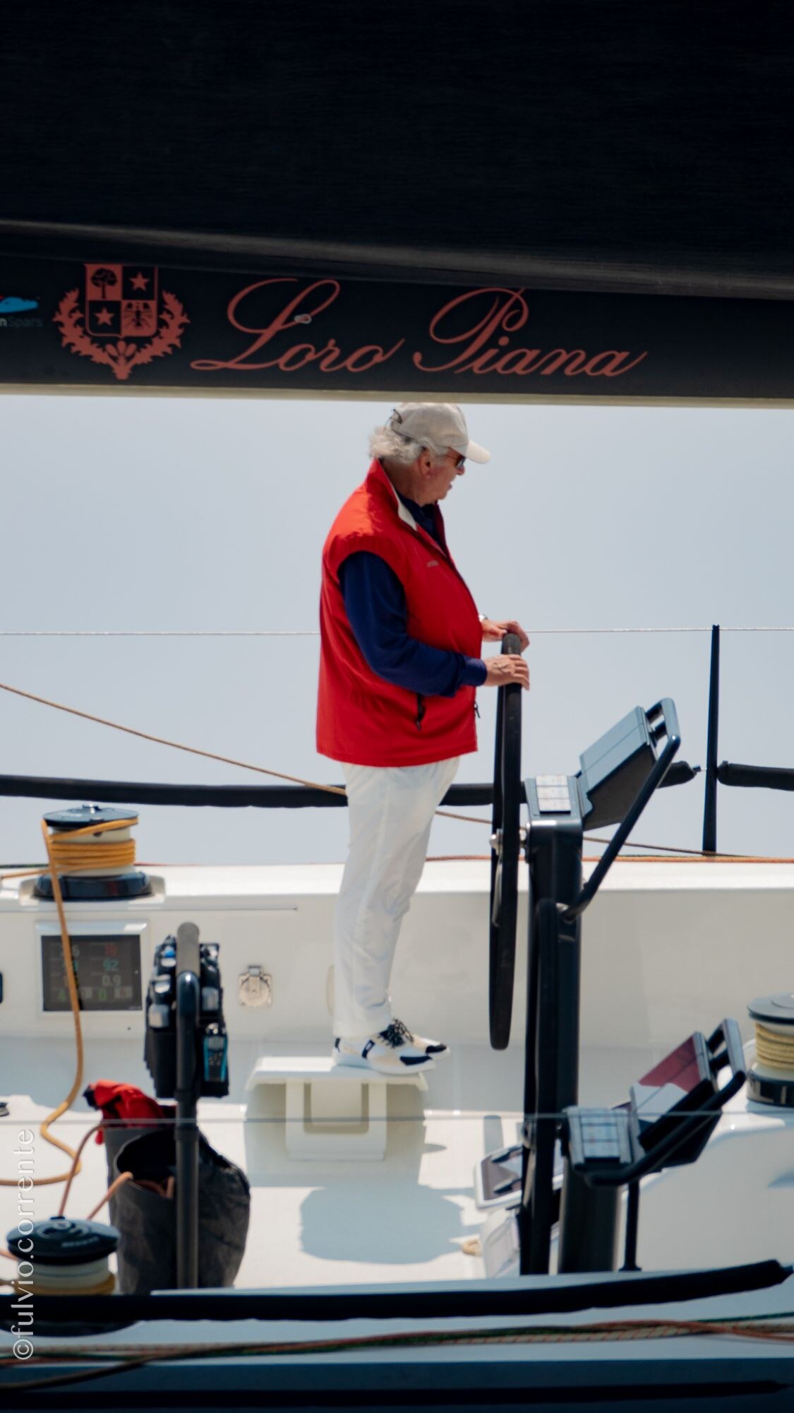 Barreur en veste rouge debout à la barre d’un voilier de course.