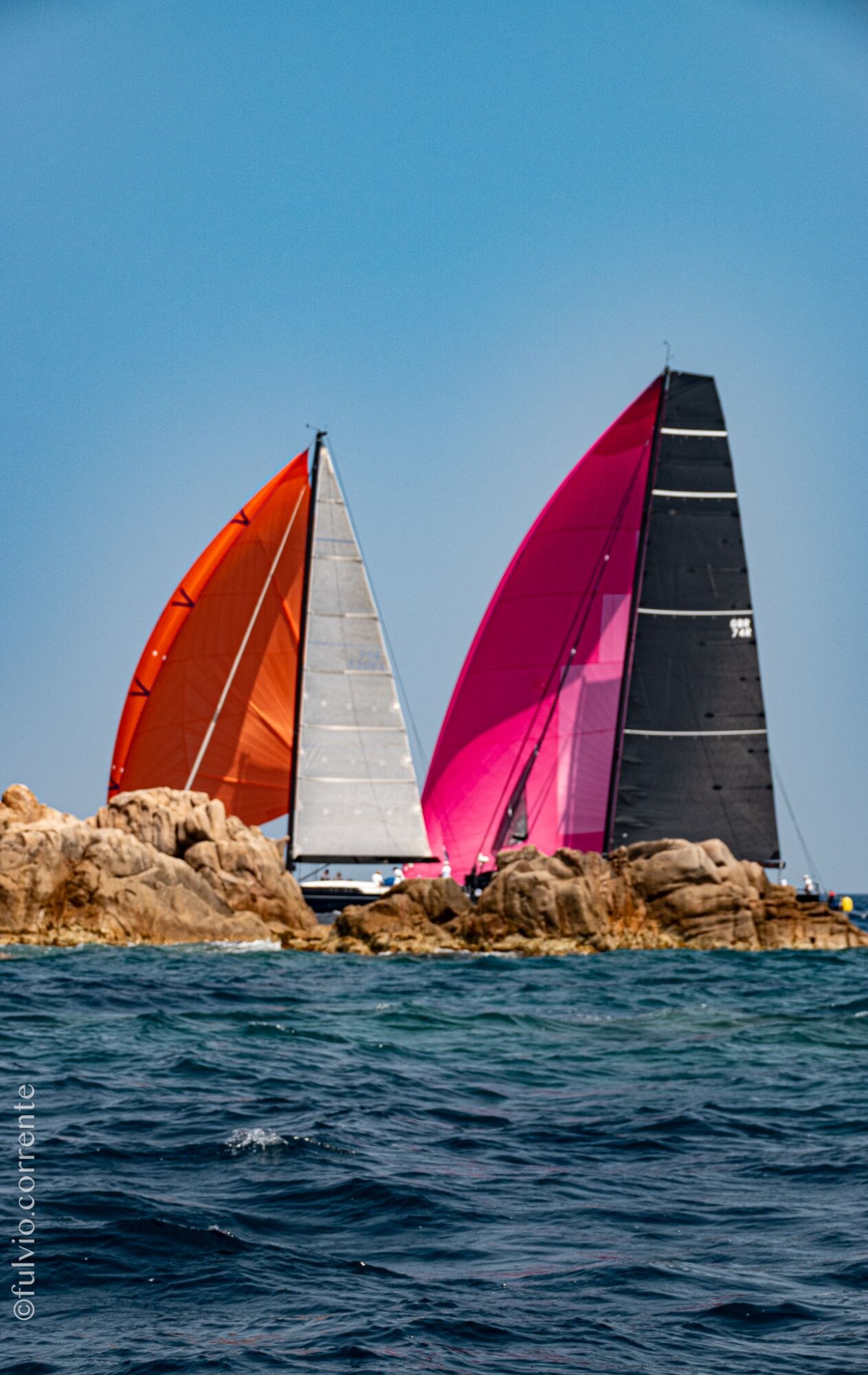Deux voiliers aux spinnakers orange et rose devant des rochers.