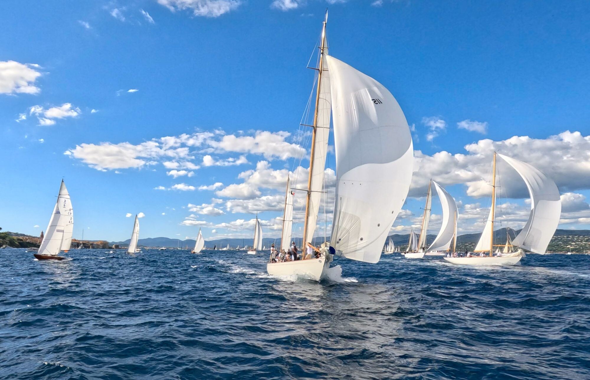Voiliers classiques aux voiles blanches, dont un spinnaker gonflé, sur une mer bleue sous un ciel de nuages.