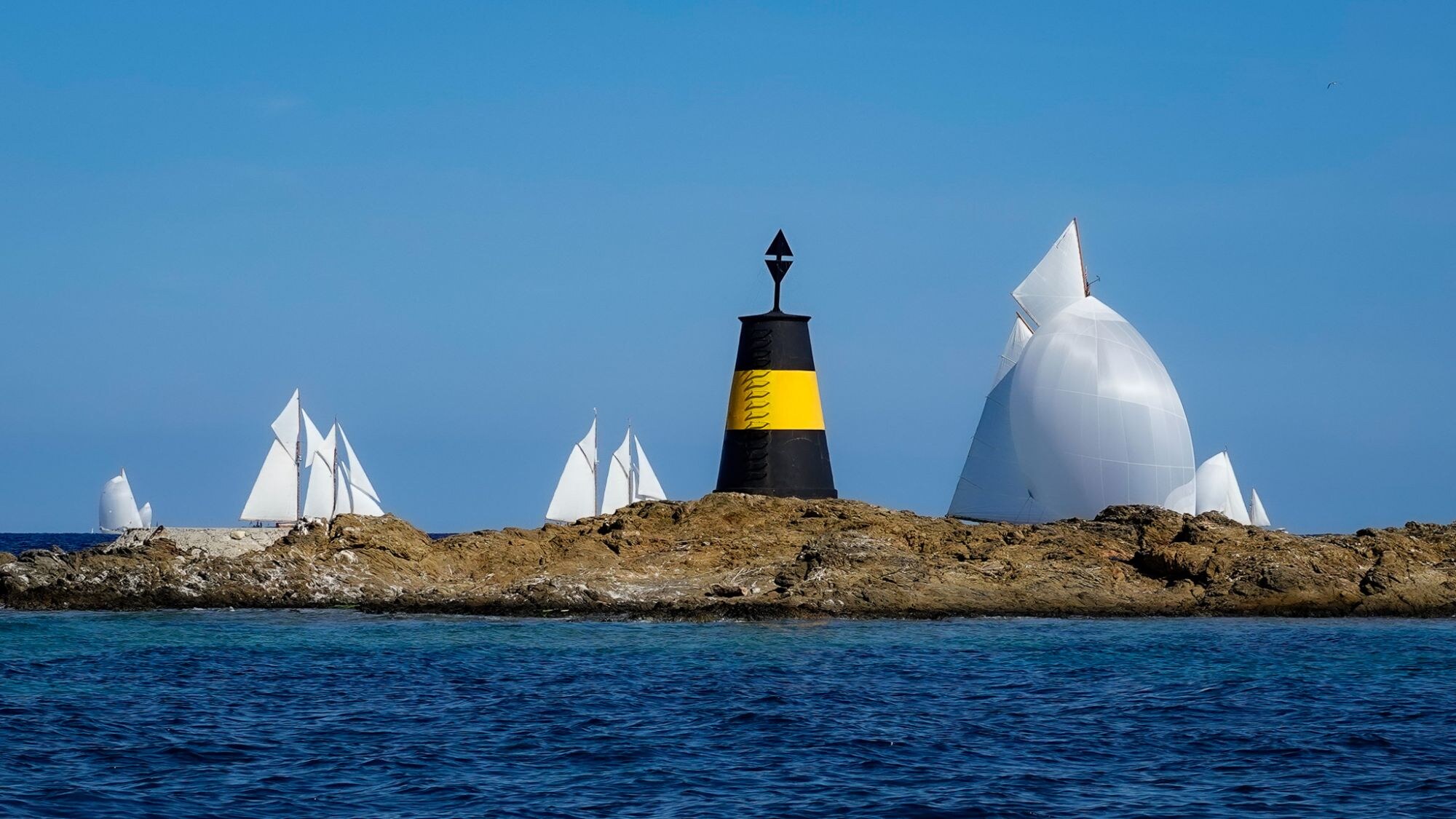 Bouée jaune et noire sur un îlot rocheux, voiliers blancs au fond sur une mer bleue.