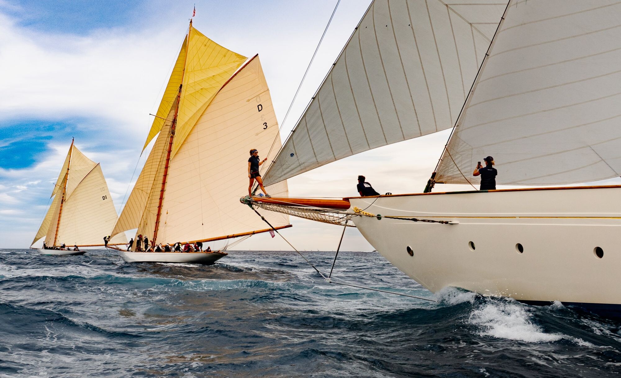 Équipiers sur l’étrave d’un grand voilier blanc, voile jaune au loin.