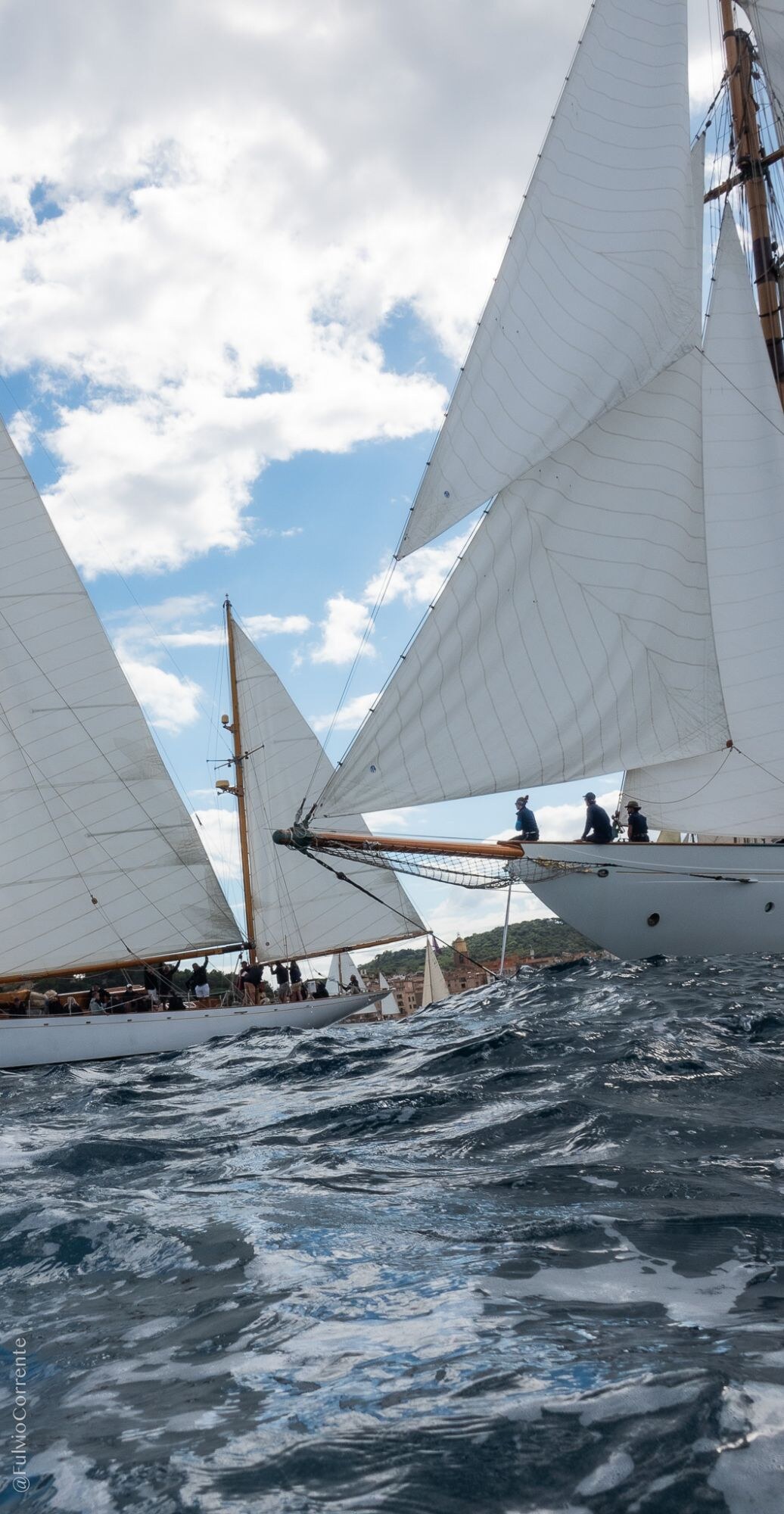 Proue d’un voilier blanc avec ses marins, vagues et écume au premier plan.