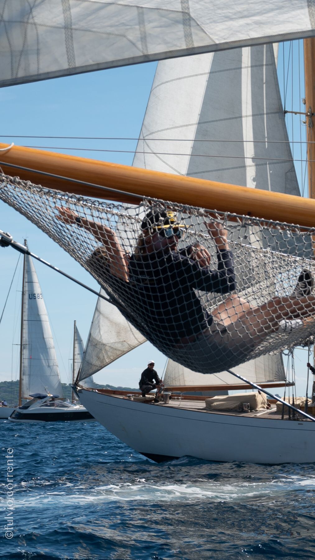 Équipier allongé dans le filet sous le bout-dehors d’un voilier classique, mer et voiles en arrière-plan.