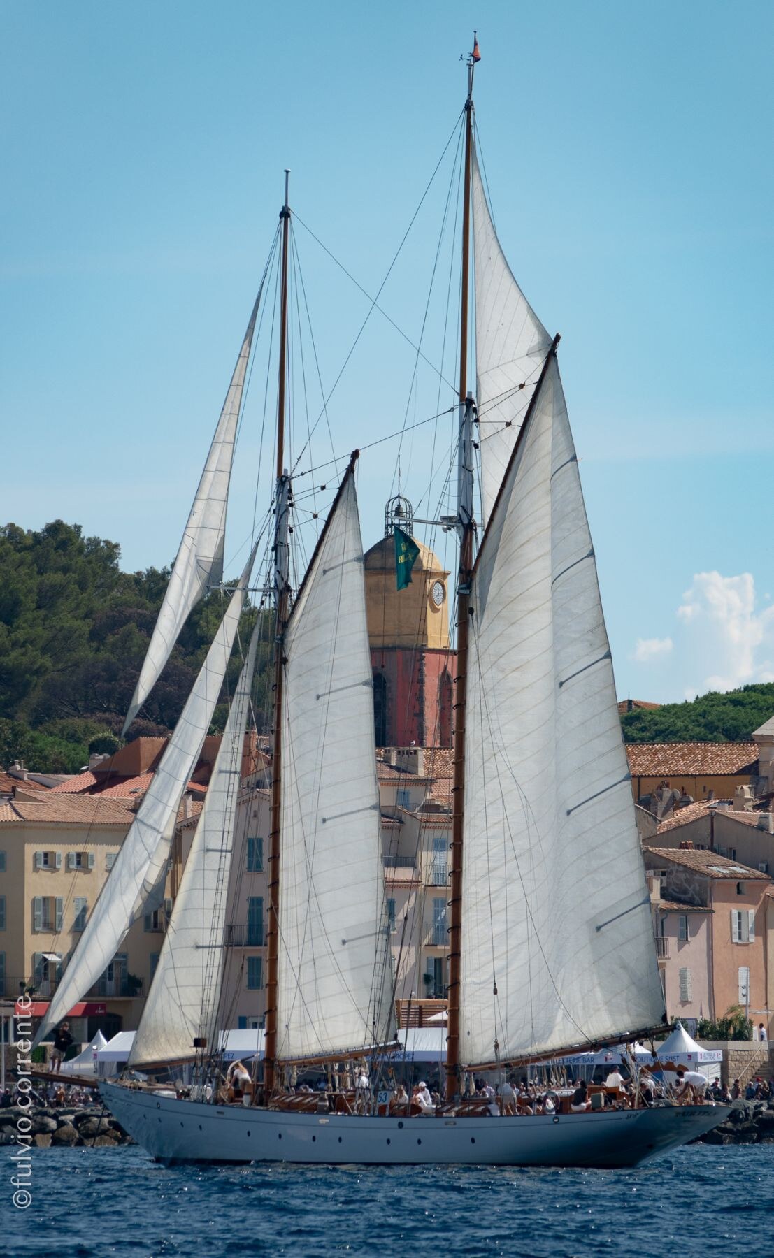 Goélette à deux mâts sous voiles devant le village de Saint-Tropez et son clocher.