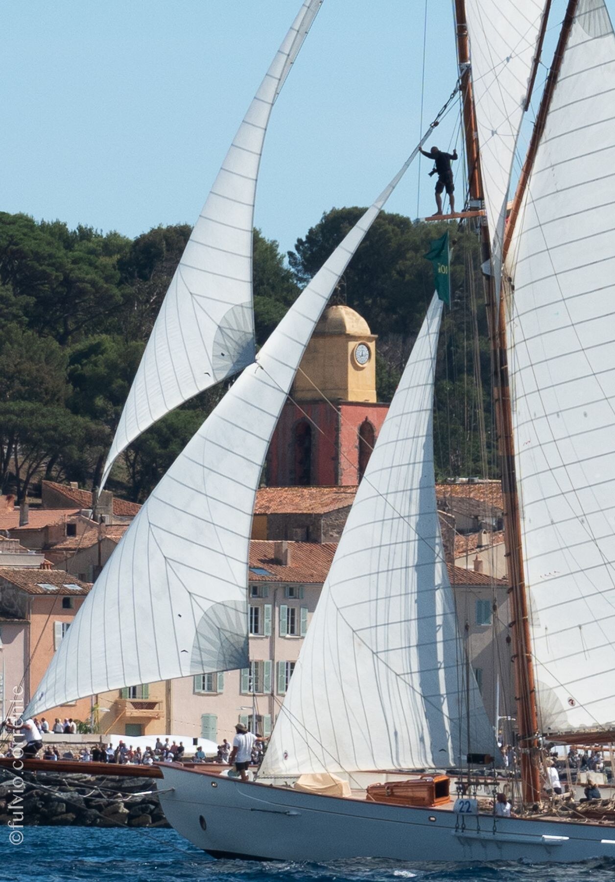 Grand voilier aux voiles gonflées devant le clocher de Saint-Tropez, un marin dans la mâture.