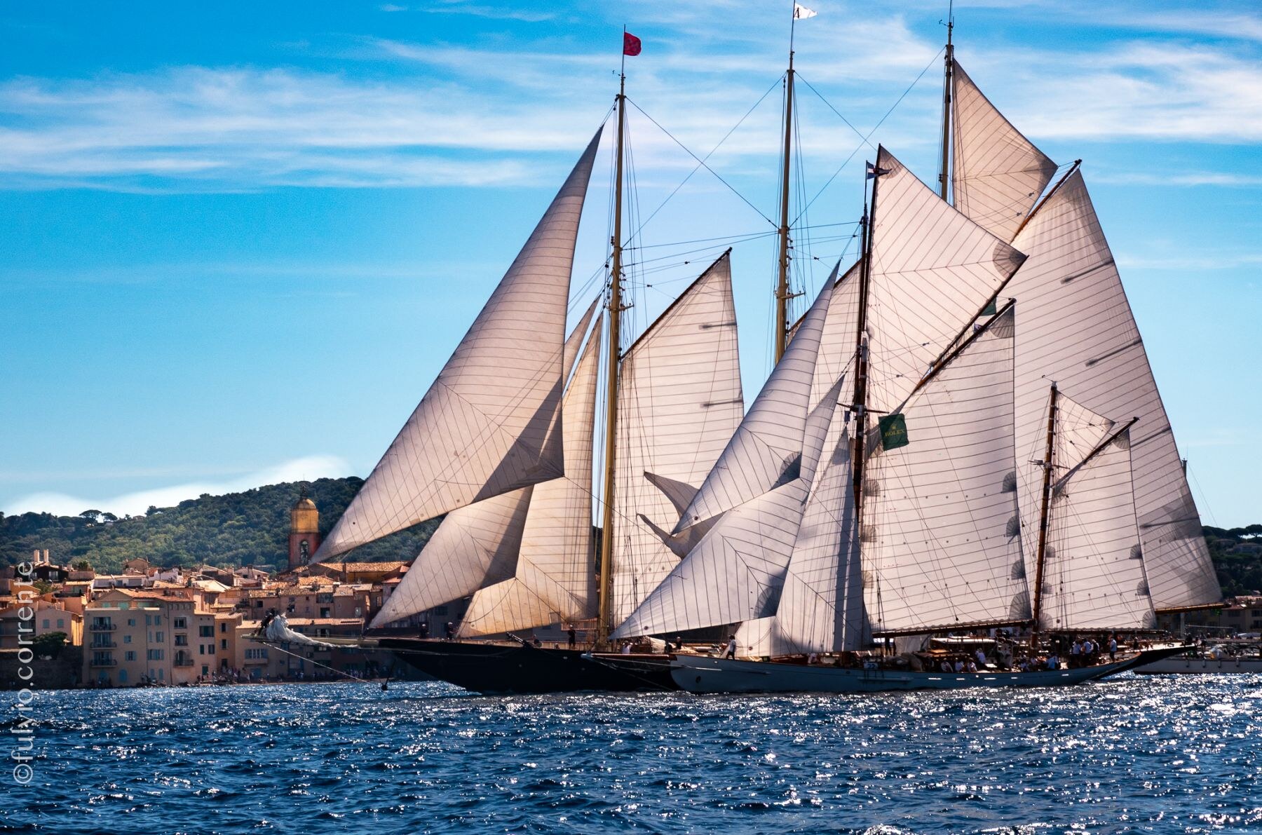 Deux goélettes aux voiles blanches se croisant sur une mer bleue devant le village.