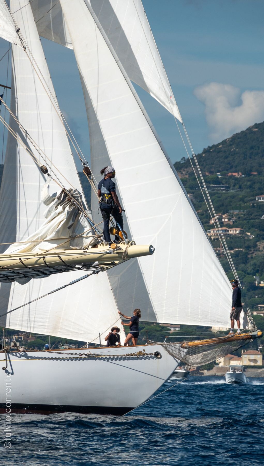 Équipiers sur le pont et le bout-dehors d’un voilier blanc, côte provençale en arrière-plan.