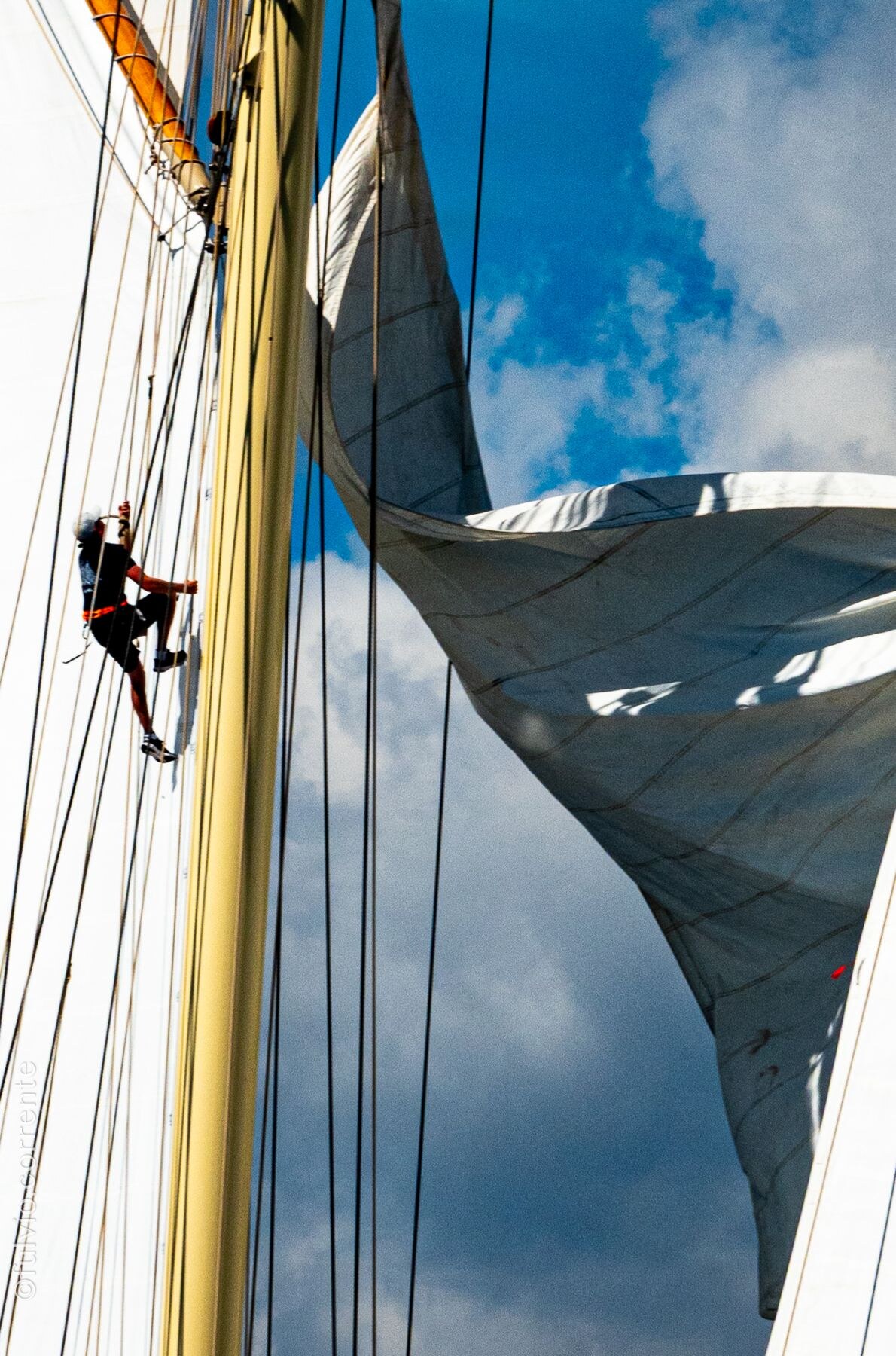 Marin grimpant le long d’un mât jaune, grande voile gonflée sur un ciel nuageux.