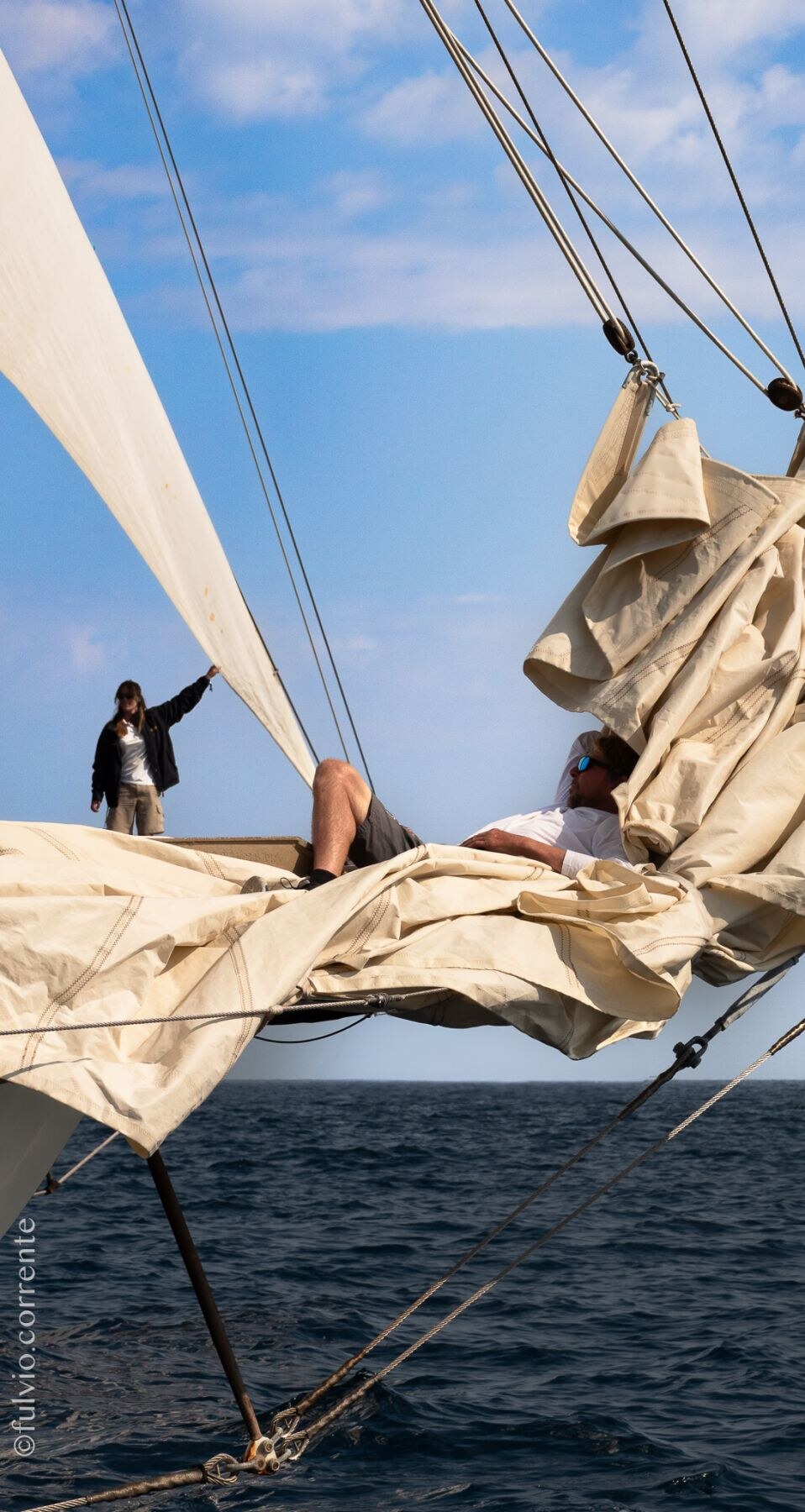 Deux équipiers sur le bout-dehors, dans les voiles beiges repliées d’un voilier.