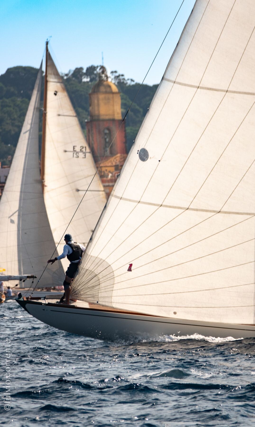 Voilier classique penché, un équipier à l’avant, devant le clocher de Saint-Tropez.