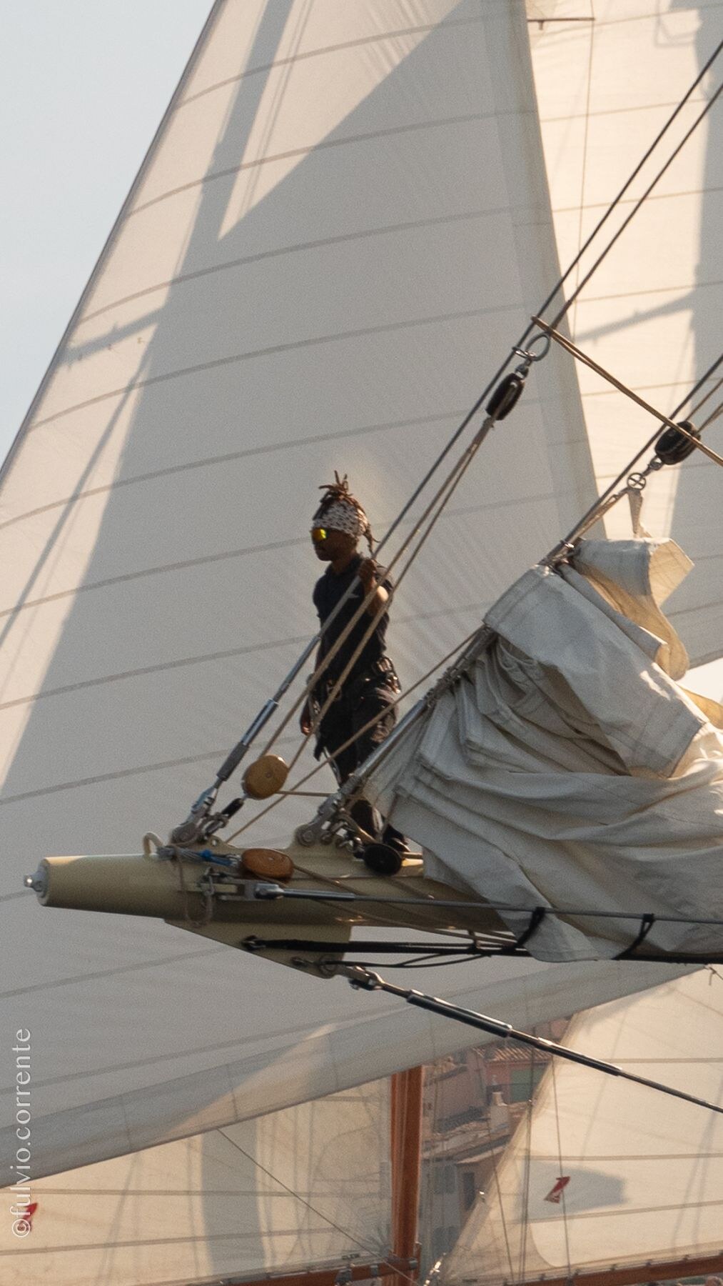 Marin debout sur le bout-dehors d’un voilier, grande voile éclairée par une lumière dorée.