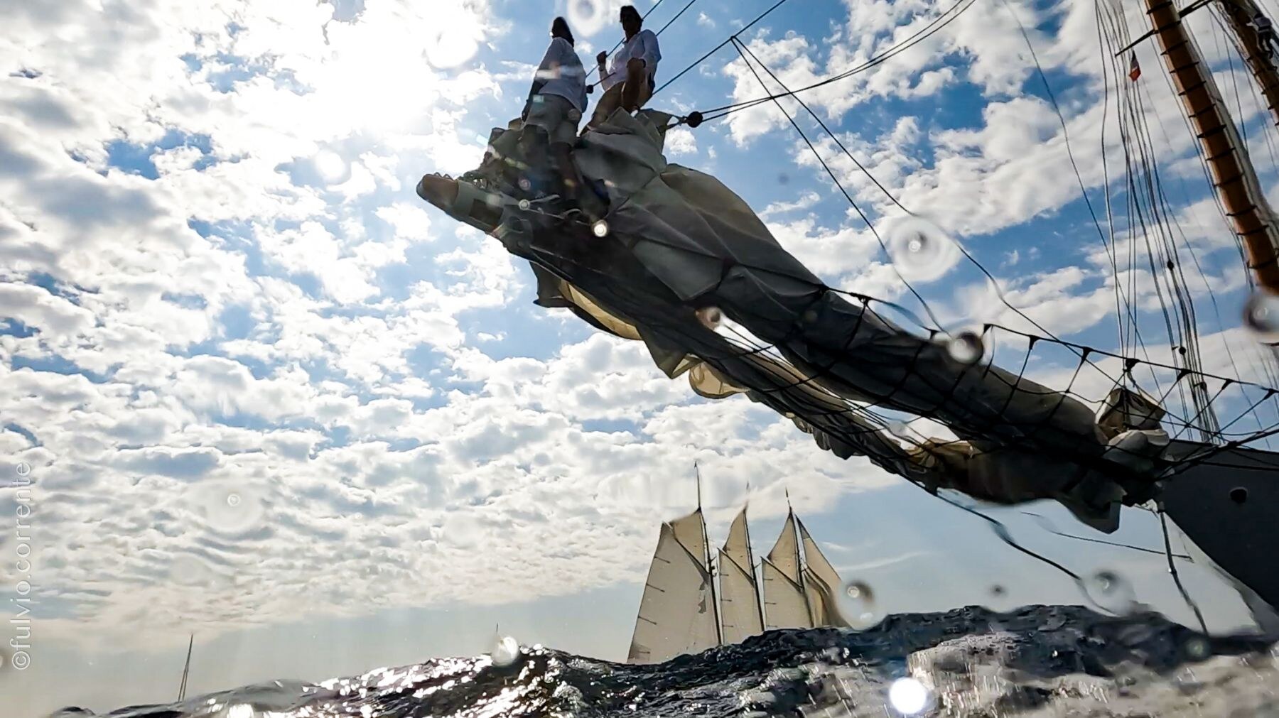 Bout-dehors d’un voilier vu en contre-plongée avec deux marins, sous un ciel moutonné.