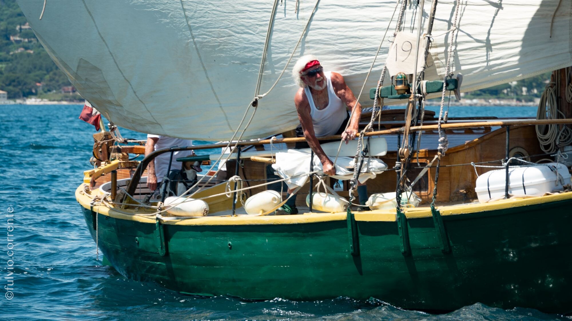 Barque à coque verte et jaune sous voile blanche, un équipier à bonnet rouge sur le pont.
