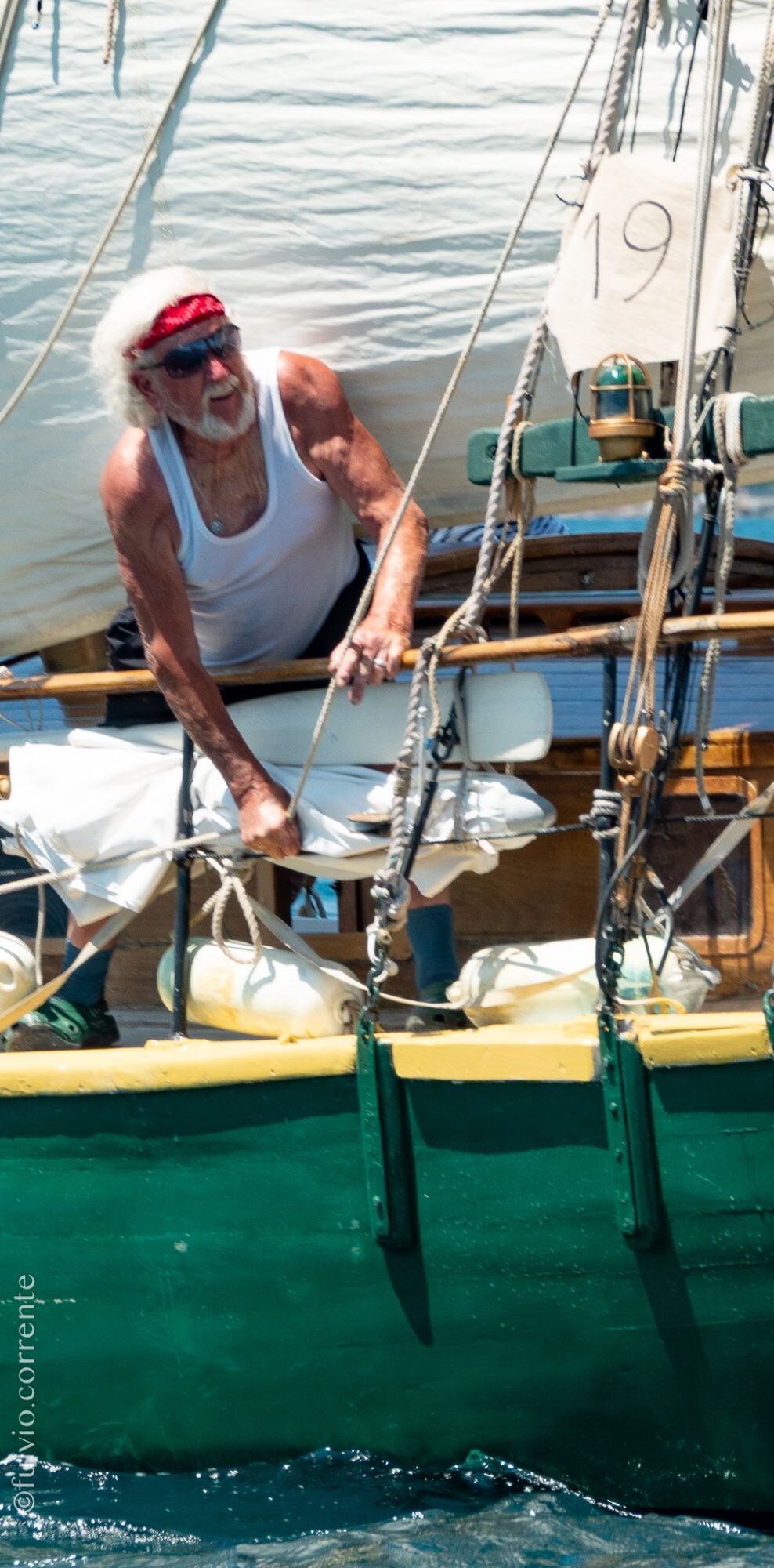 Équipier aux cheveux blancs et bandeau rouge penché au-dessus du bastingage d’une barque verte, numéro 19 sur la voile.